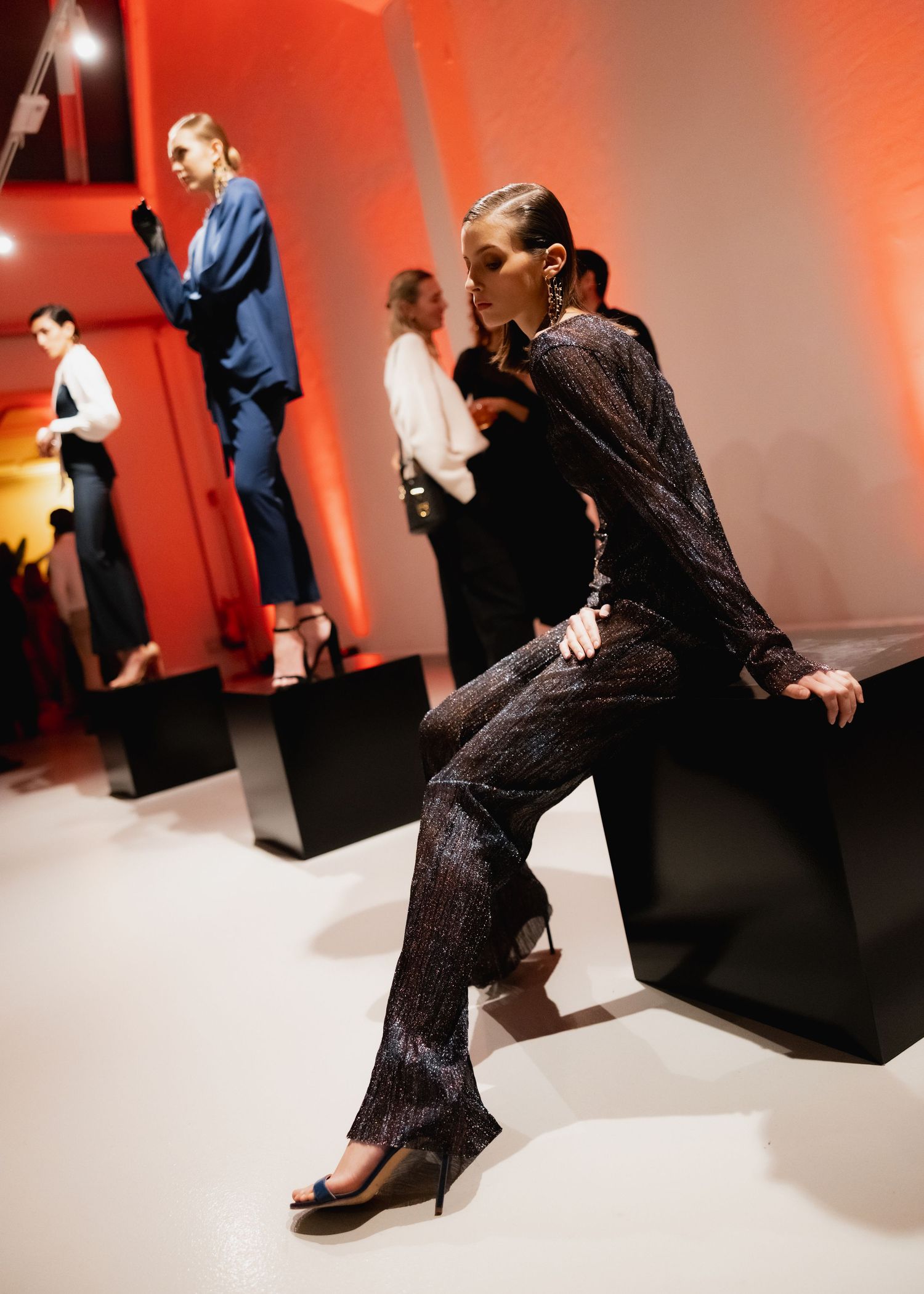 Models pose on geometric black blocks against a red-orange wall during a fashion presentation.