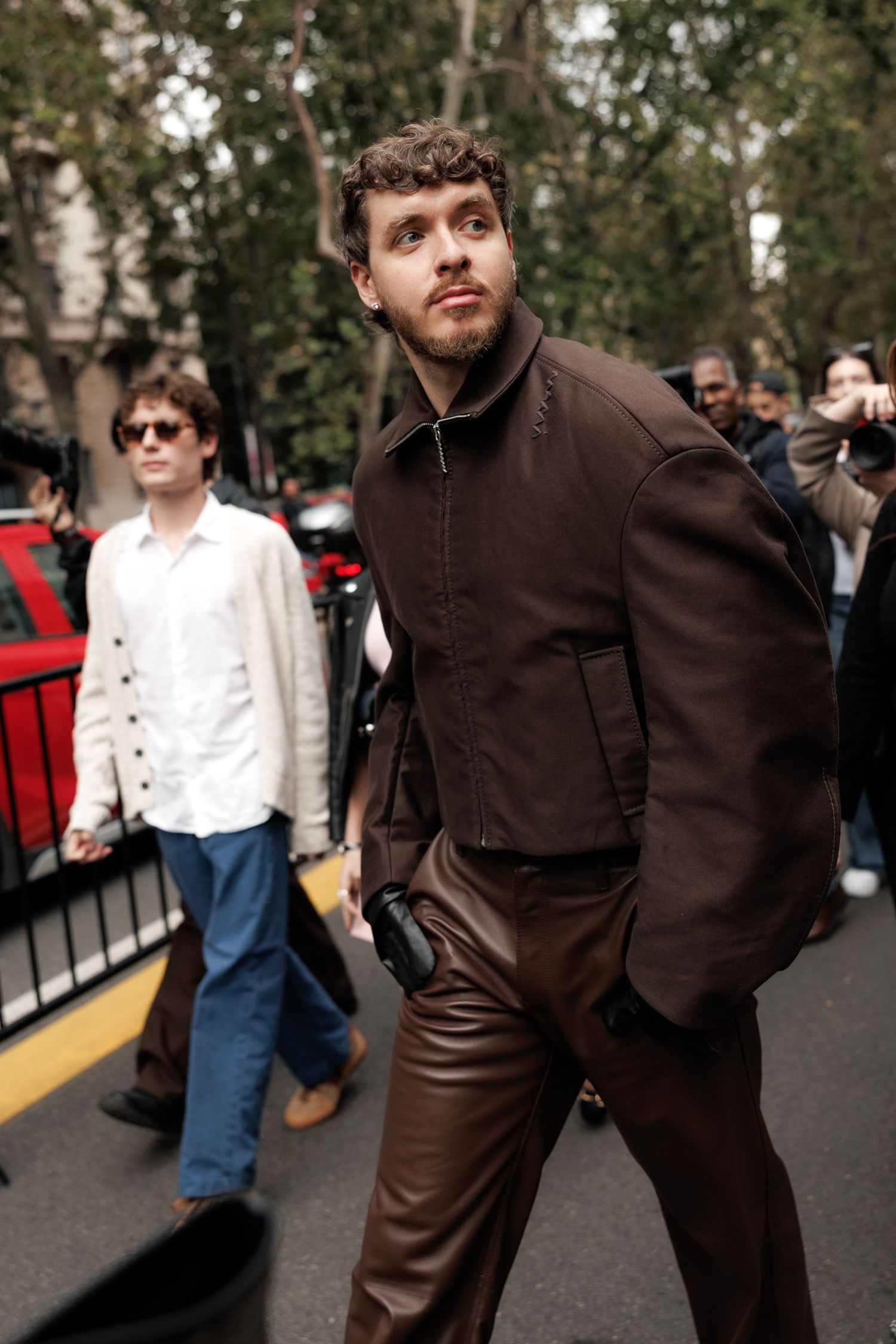 Street fashion photo showing person in brown leather pants and dark jacket with casual crowd in background.