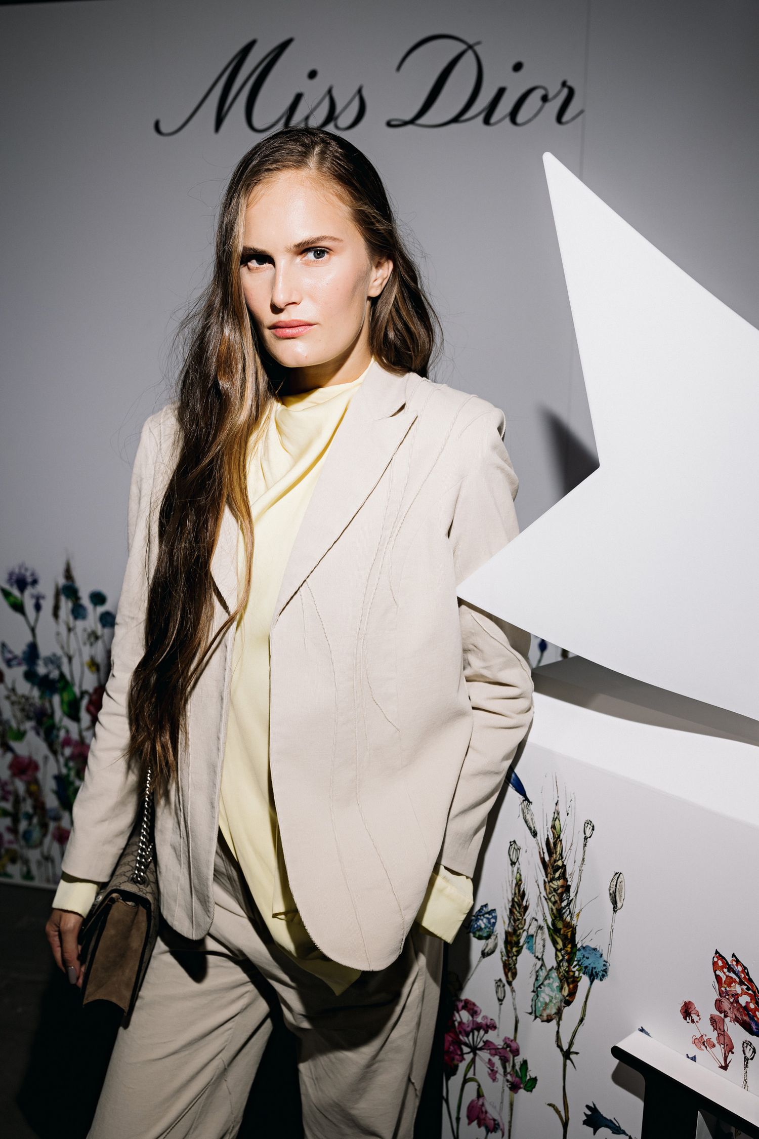 Fashion model in beige blazer and pants poses against Miss Dior branded wall with floral decorations.