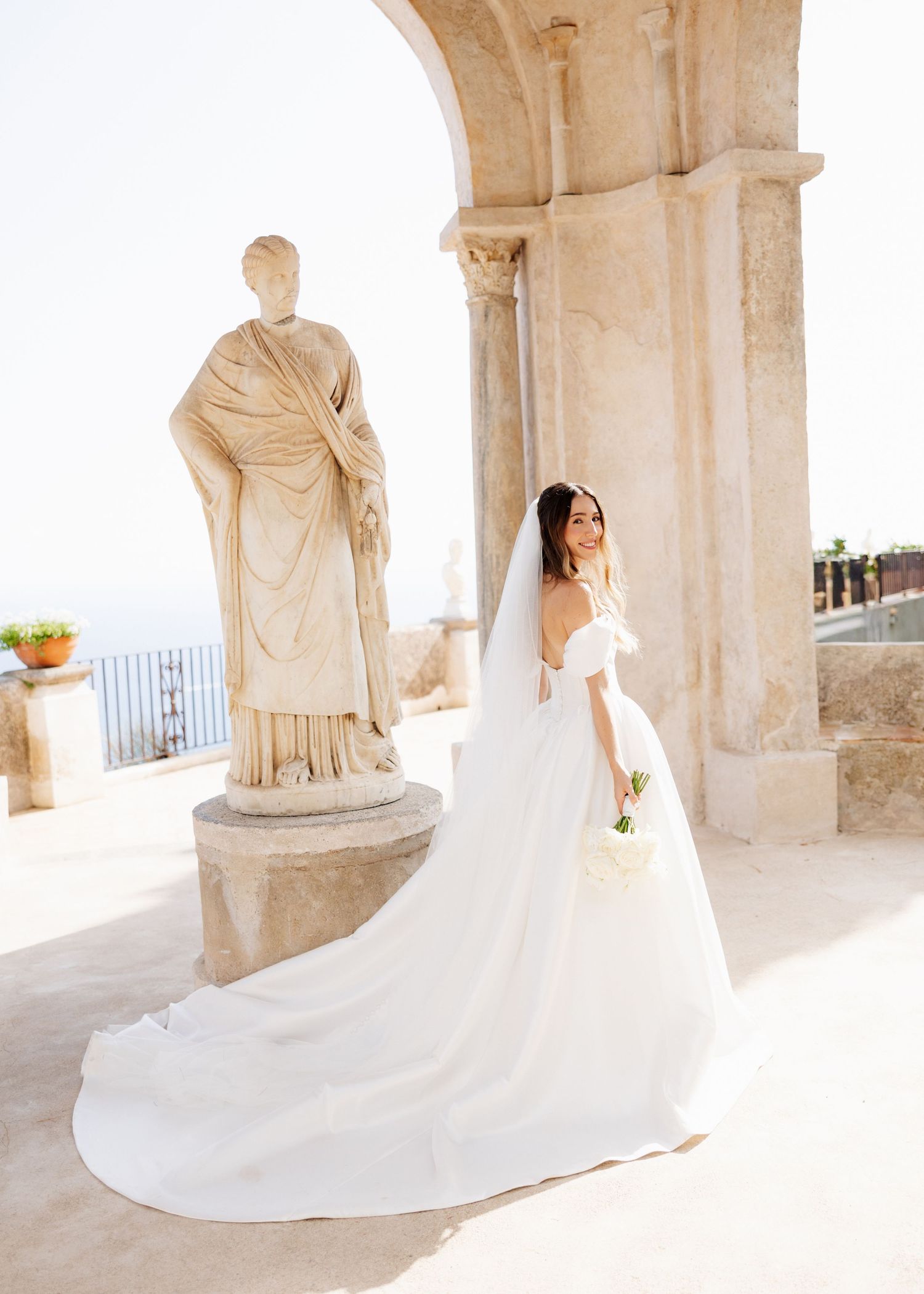 Bride in off-shoulder gown with cathedral veil poses near classical statue on sunlit Italian terrace.