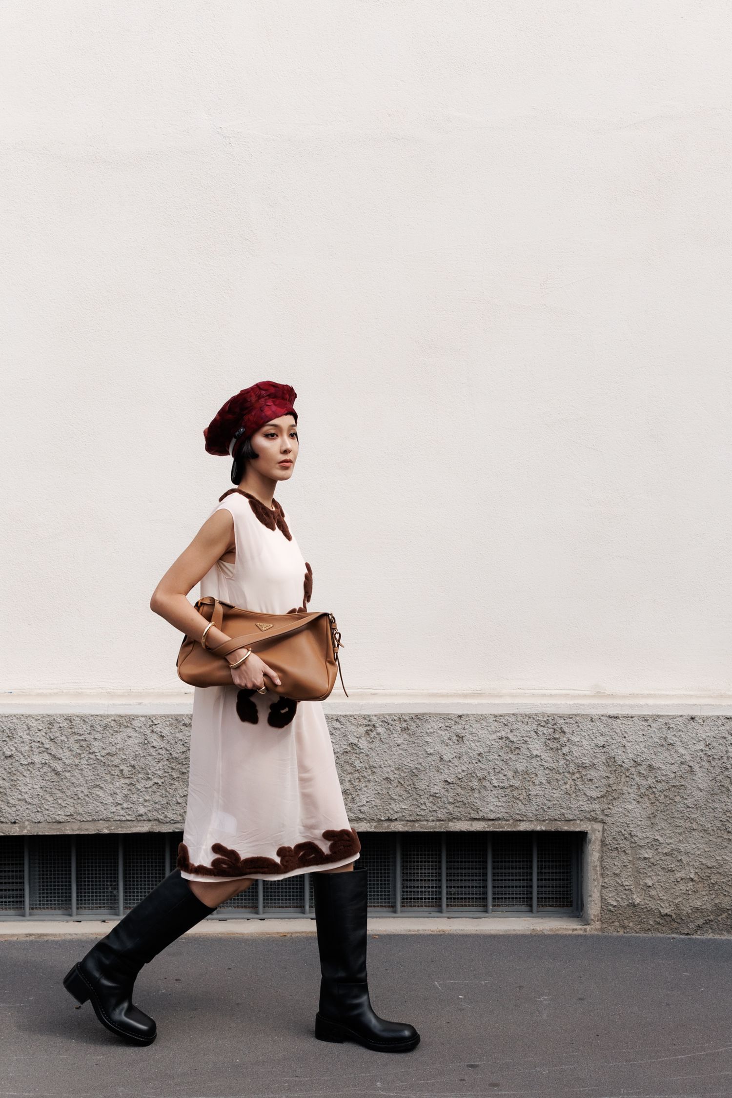 Woman in vintage style dress and red beret walking past grey wall with leather bag.