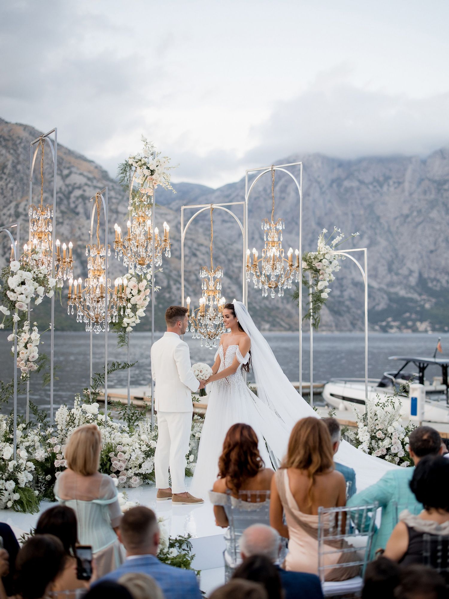 Elegant lakeside wedding ceremony with crystal chandeliers and white floral arrangements against mountain backdrop.