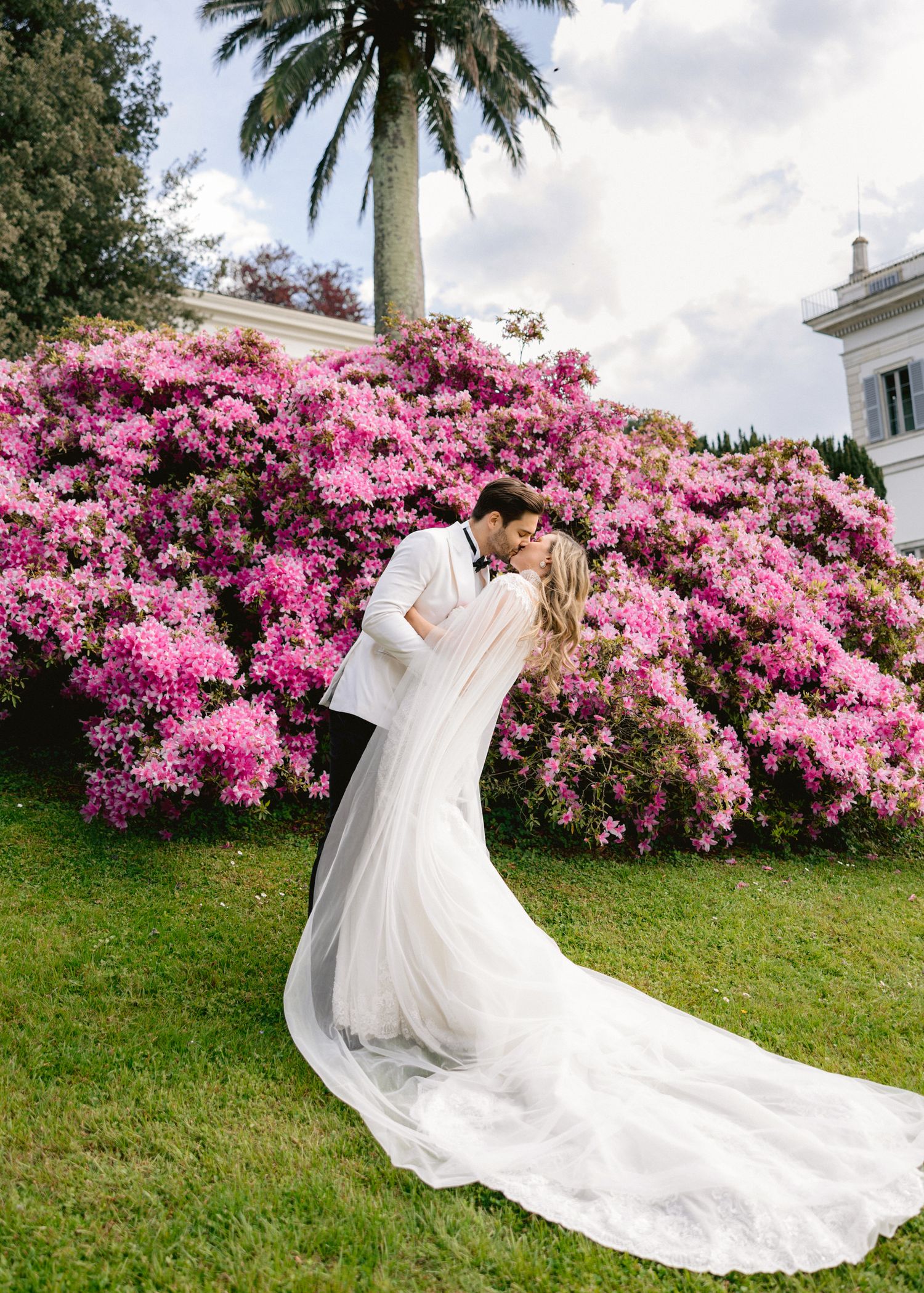 Bride and groom share a romantic kiss in front of vibrant pink azalea bushes on lush green lawn.