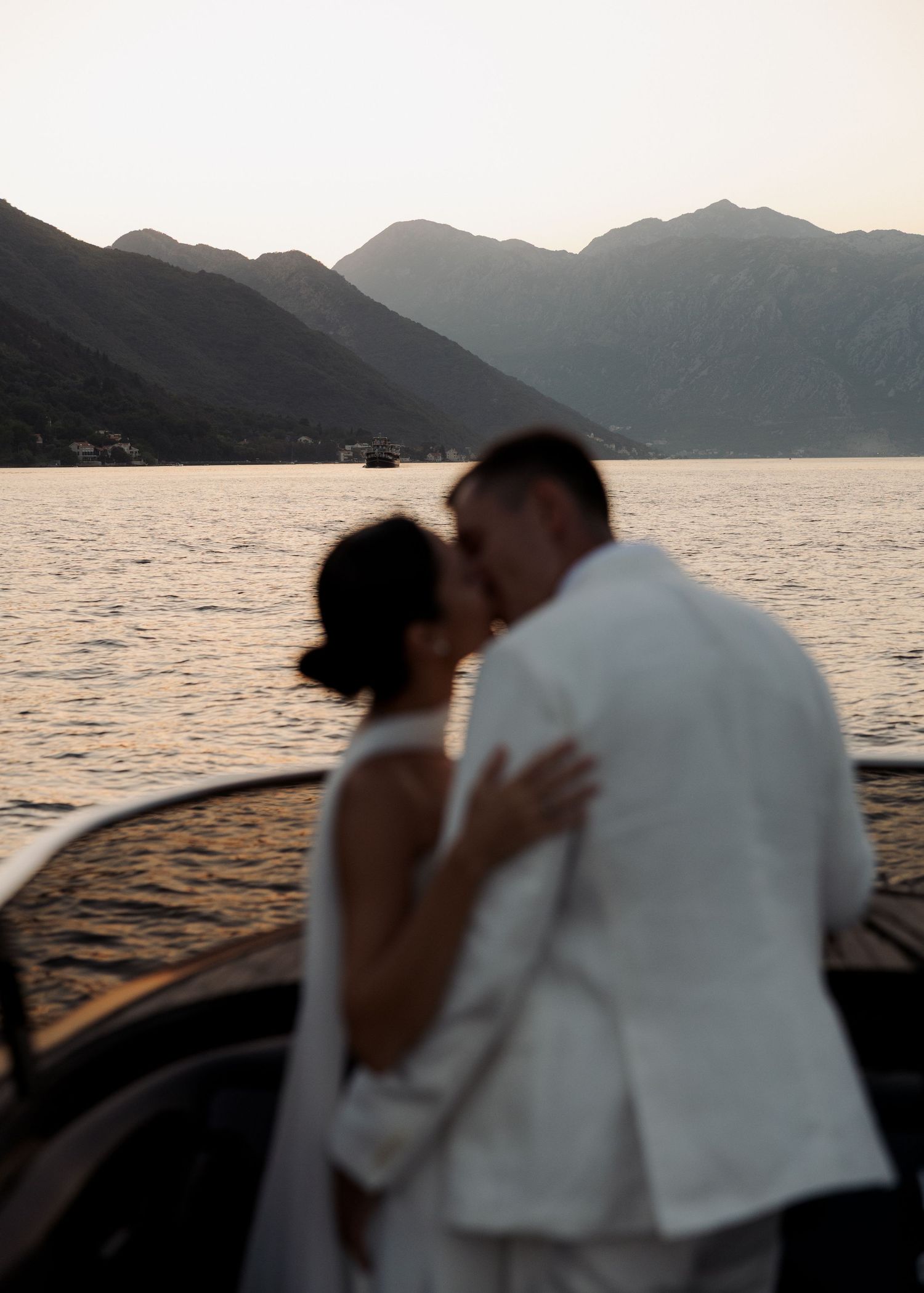Romantic silhouettes embrace on a boat at sunset with mountains and lake in the background.