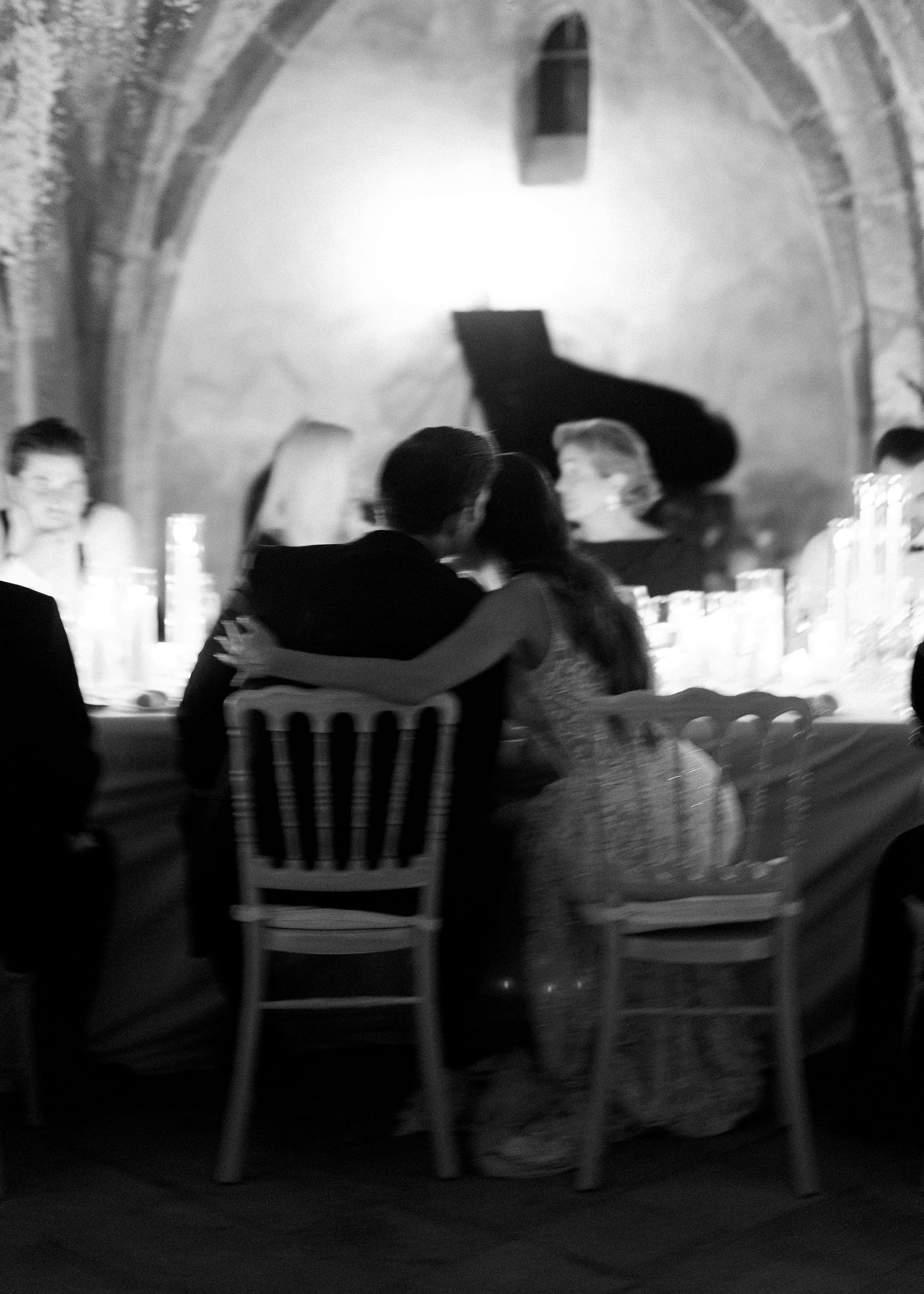 Black and white photo of a couple sharing an intimate moment at a candlelit dinner table in an elegant stone-arched venue.