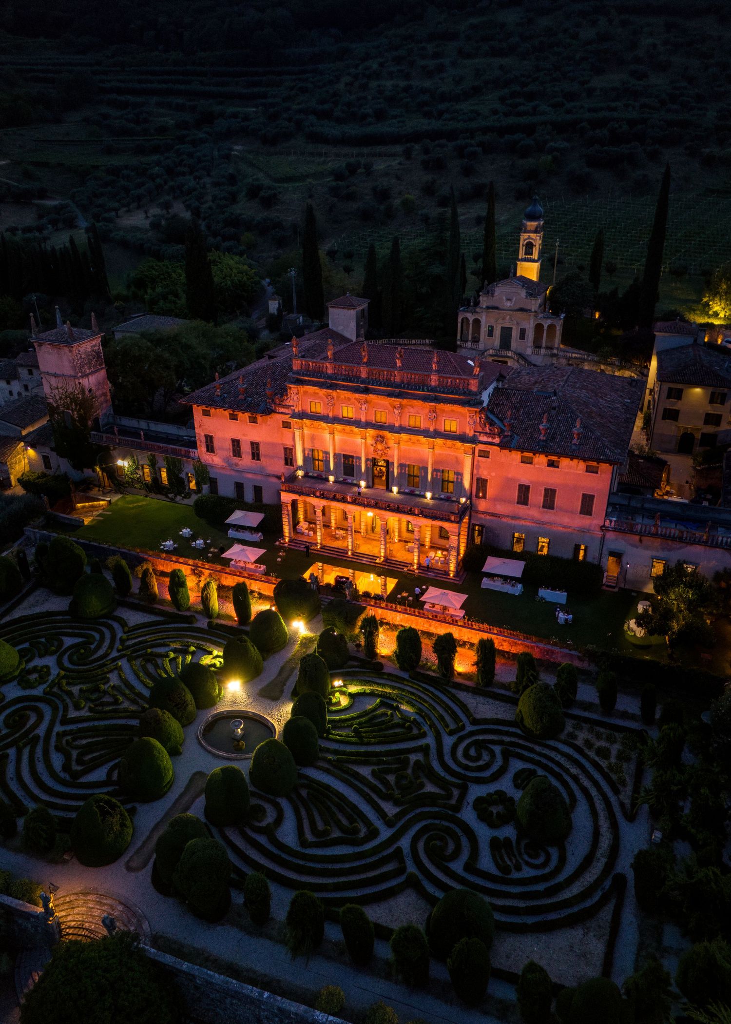 Aerial night view of an illuminated Italian villa with ornate formal gardens, glowing orange against the dark landscape.
