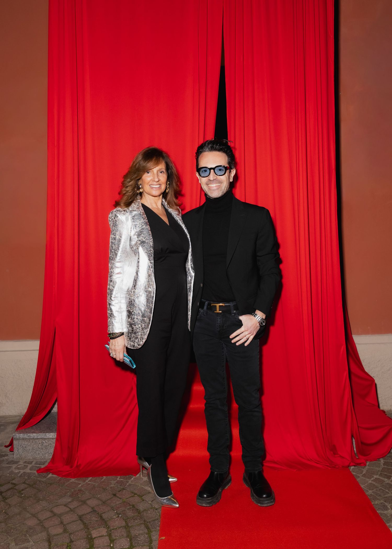 Two people pose together against a dramatic red curtain backdrop, one wearing a sparkly silver blazer and one in all black.
