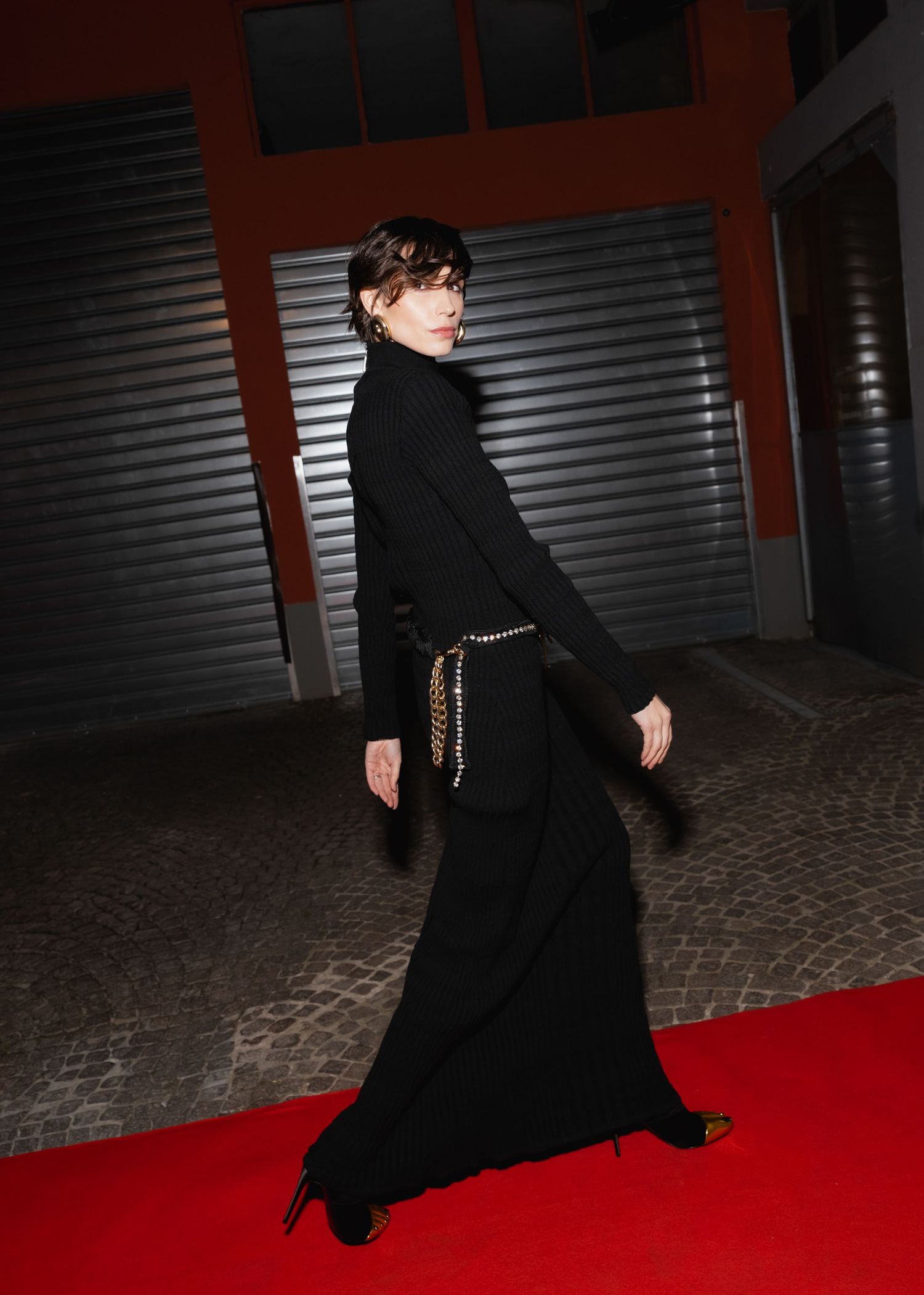 A person in a black coat and hat walks across a red carpet against a metallic backdrop in a dramatic pose.
