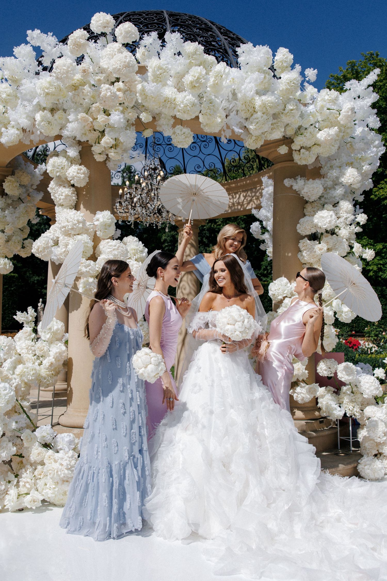 Wedding party poses under spectacular white floral arch with parasols.