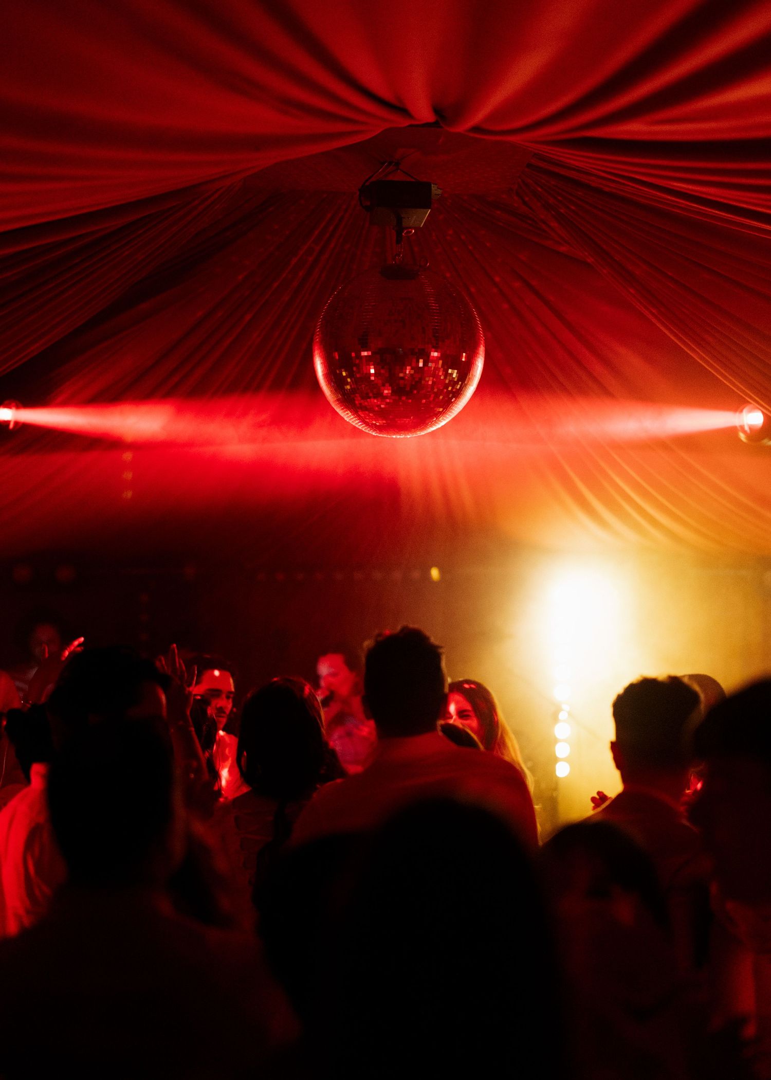 A crowded dance floor glows under a disco ball and red laser lights inside a draped event venue.