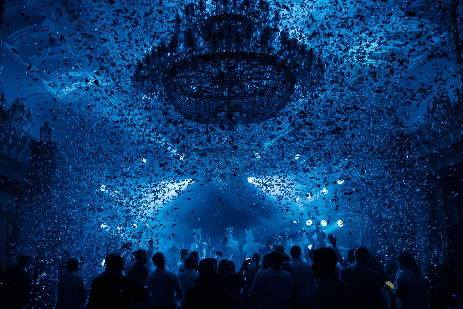A crowd silhouetted beneath a dramatic blue-lit chandelier in a dark venue during an event.