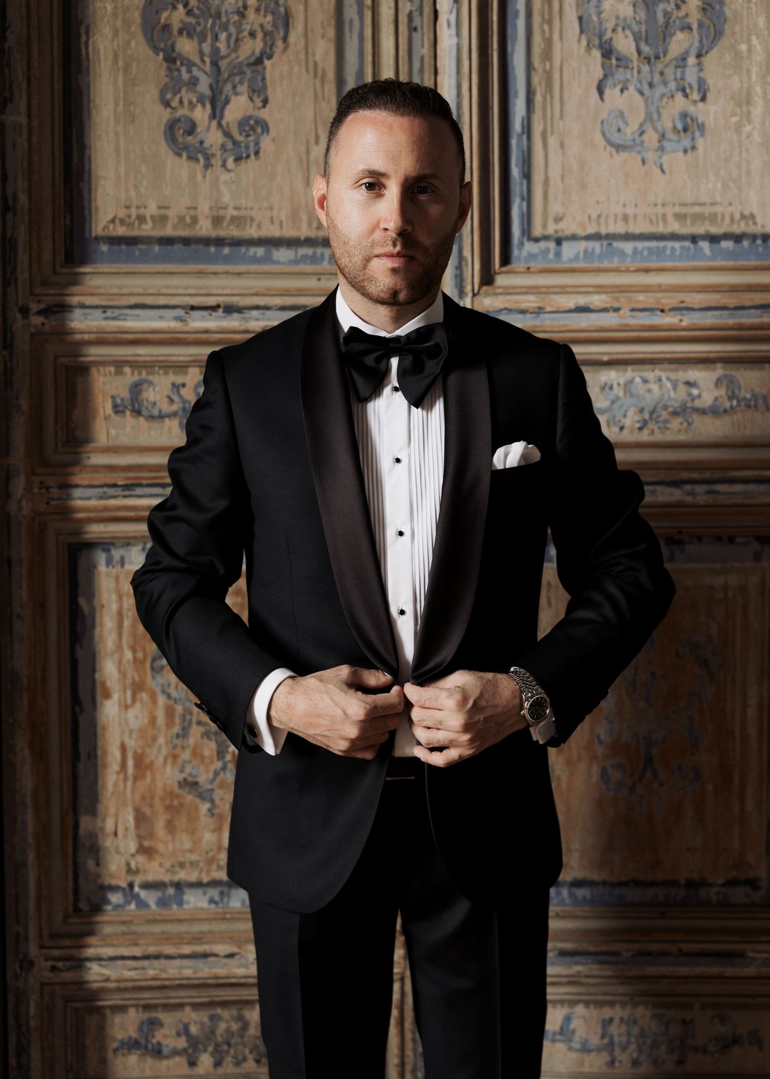 Man in black tuxedo with bow tie and white pocket square buttoning jacket in front of ornate wooden door.