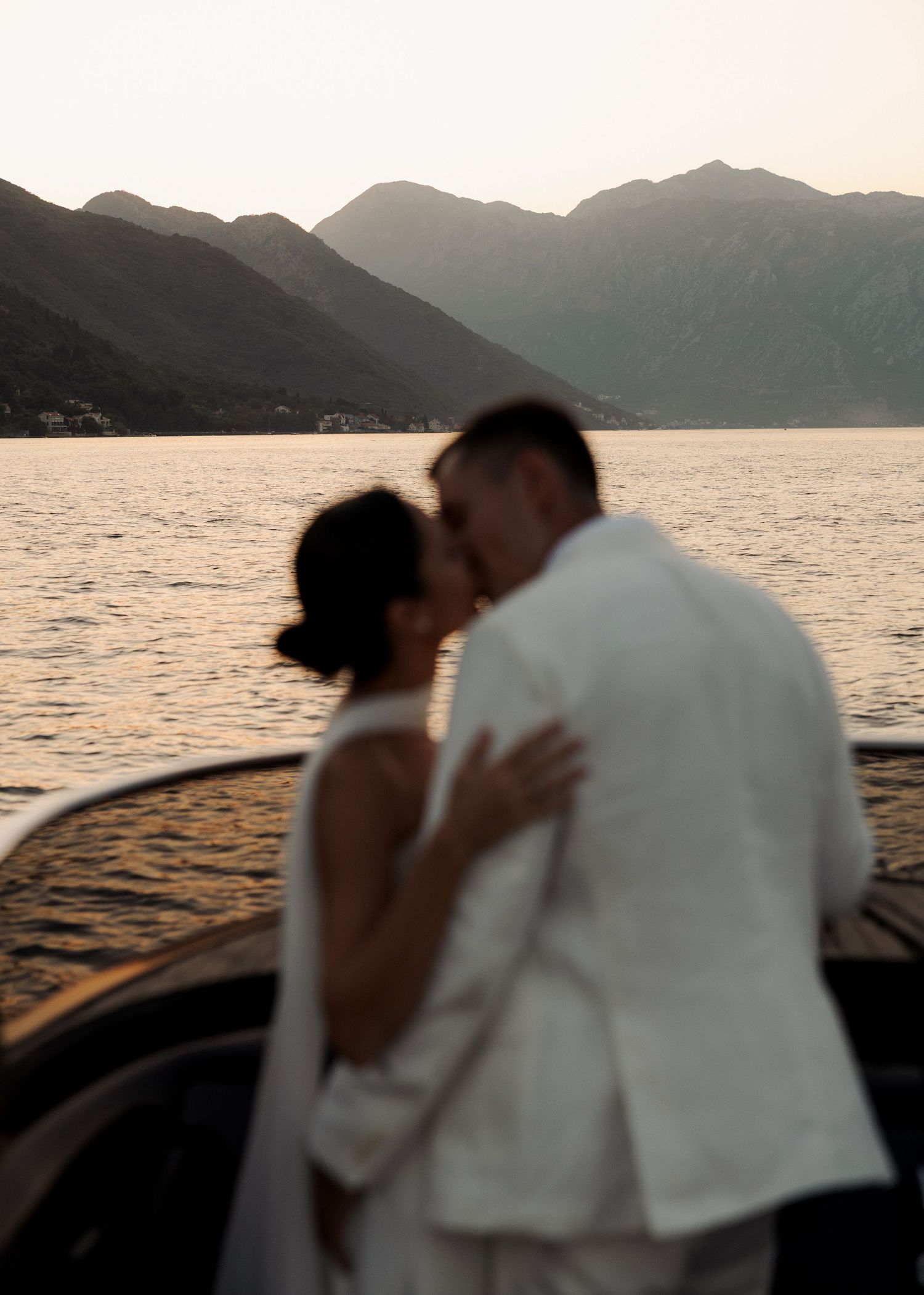 Couple in white attire sharing a romantic kiss on a boat with mountain lake backdrop at sunset.