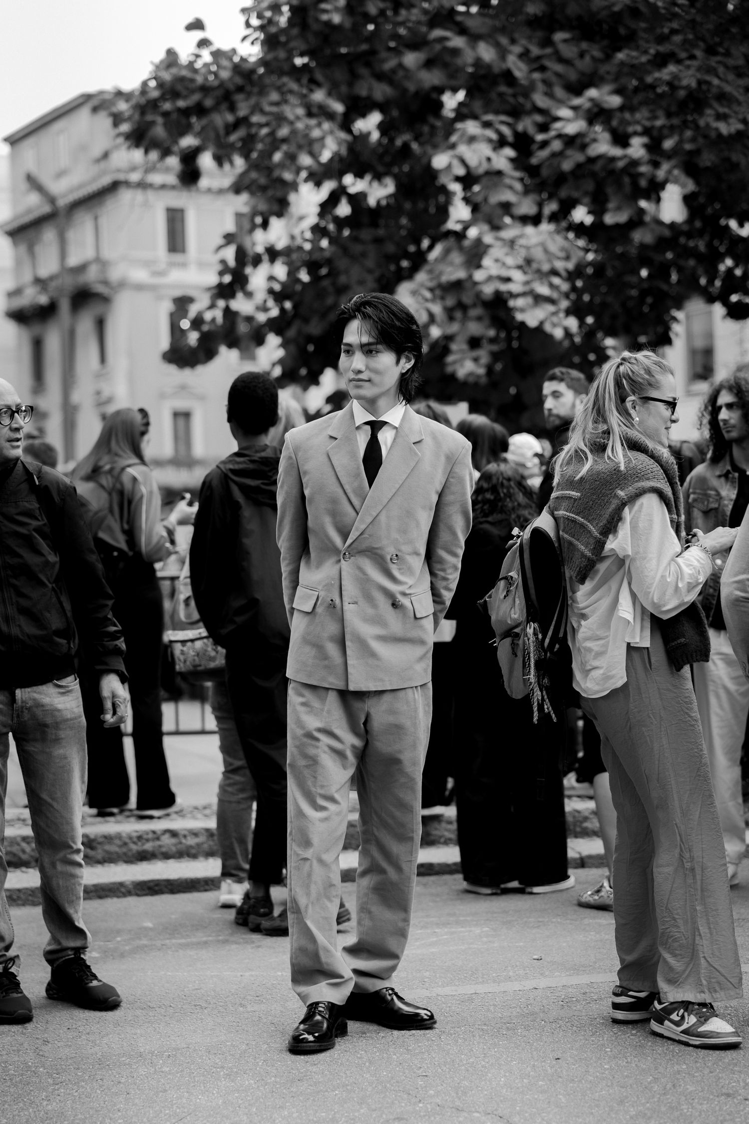 A black and white photo of someone wearing a light-colored suit standing among a crowd of people outdoors.