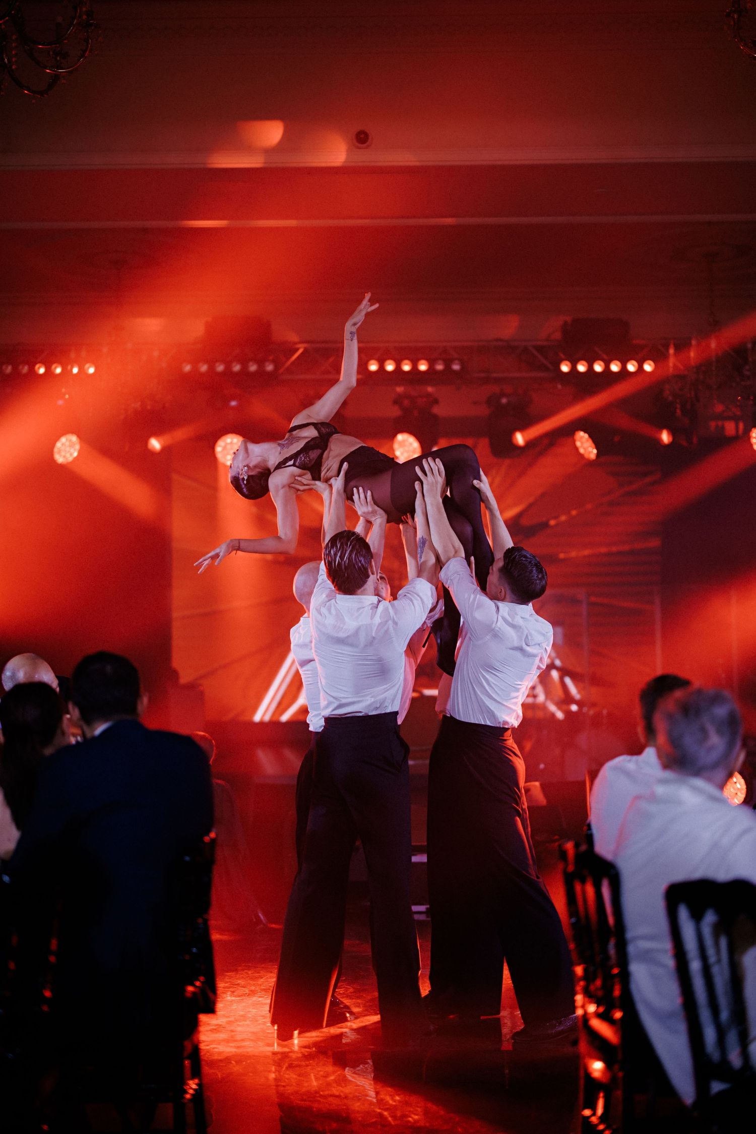 Dancers perform a dramatic lift in white outfits under dramatic red stage lighting at a theatrical event.
