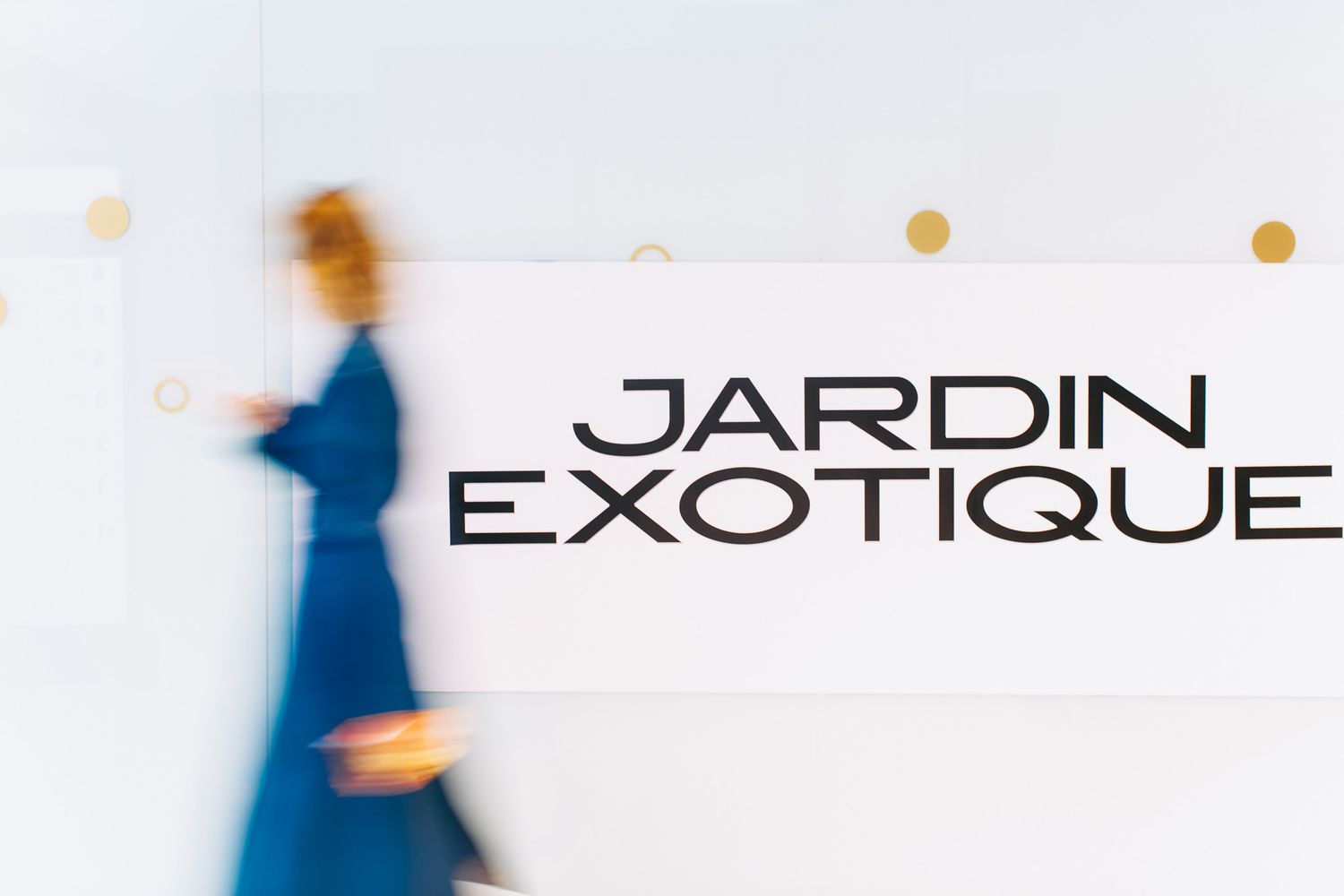 Motion-blurred figure passes by Jardin Exotique text display on white wall.