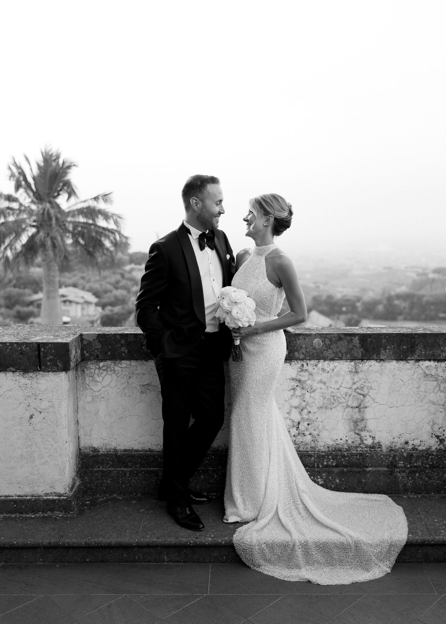 Black and white photo of a smiling bride and groom on a terrace with palm trees and scenic landscape backdrop.