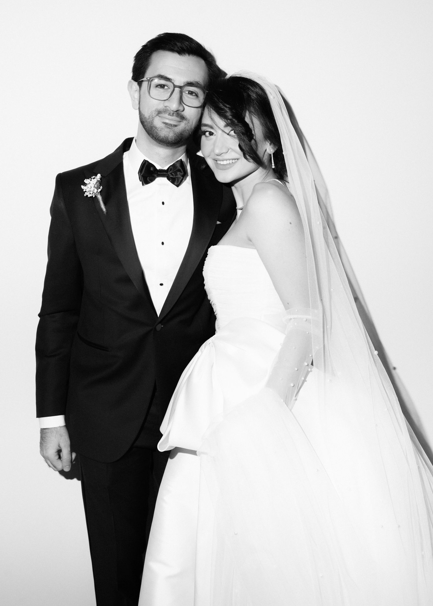 Black and white wedding portrait of bride in strapless gown with veil and groom in tuxedo with bow tie and glasses.