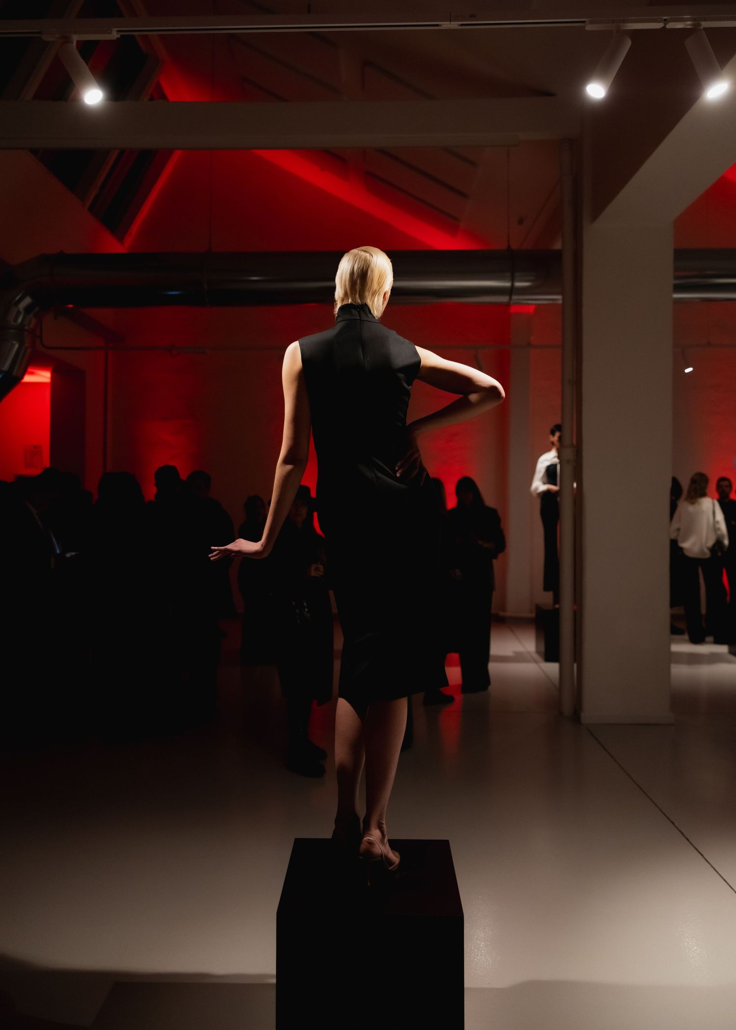 A dancer in black performs graceful movements in a dimly lit studio space with red accent lighting.