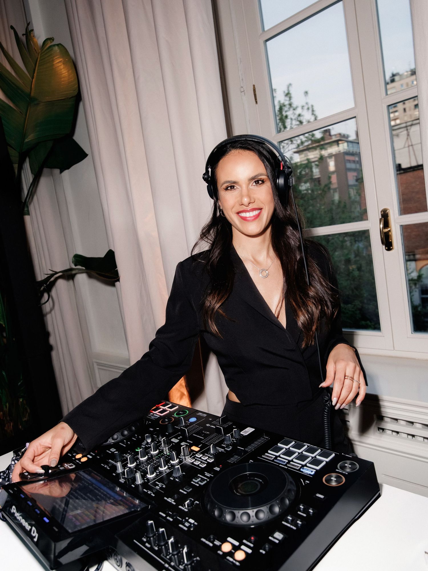 A DJ works behind professional mixing equipment at a venue with windows in the background.