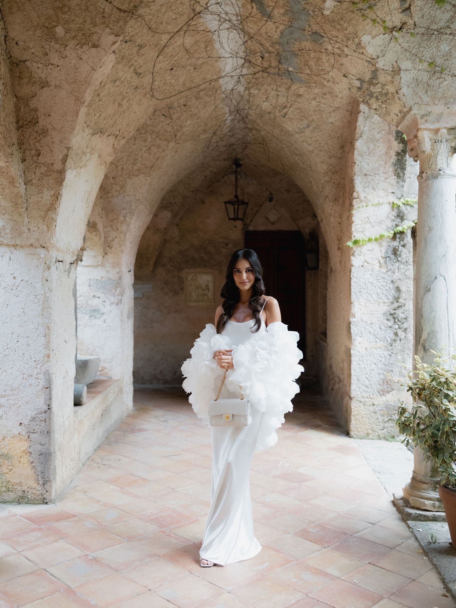 A couple in white wedding attire embraces under stone archways in a rustic Mediterranean courtyard setting.