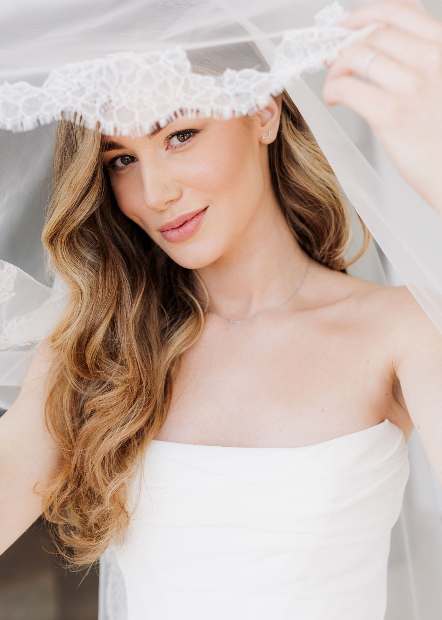 Bride in white strapless gown lifting lace-trimmed veil, with soft wavy blonde hair and natural bridal makeup.