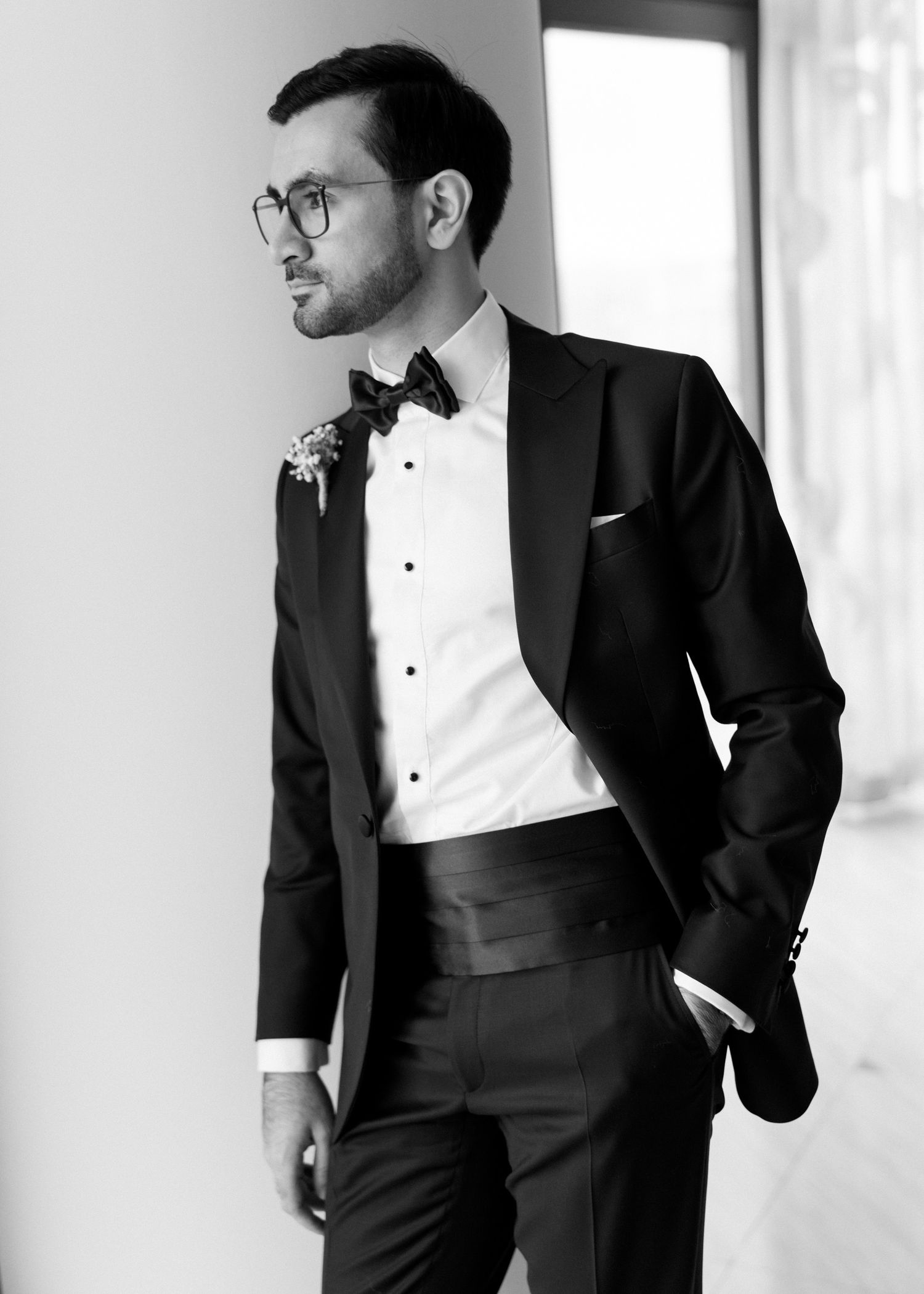 A dapper groom in a black tuxedo with bow tie and boutonniere poses confidently in a black and white portrait.