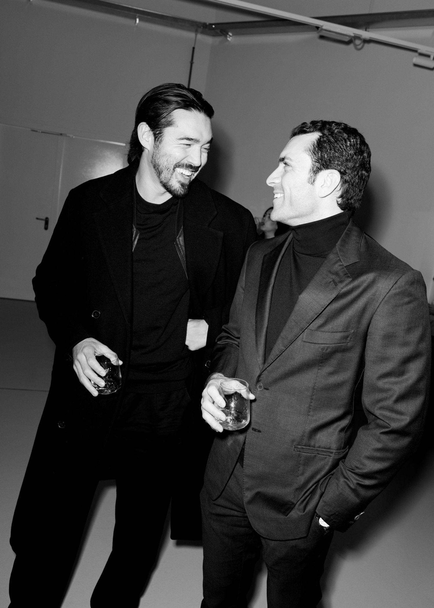 Two men in black suits share a candid moment of laughter in a black and white photo series.