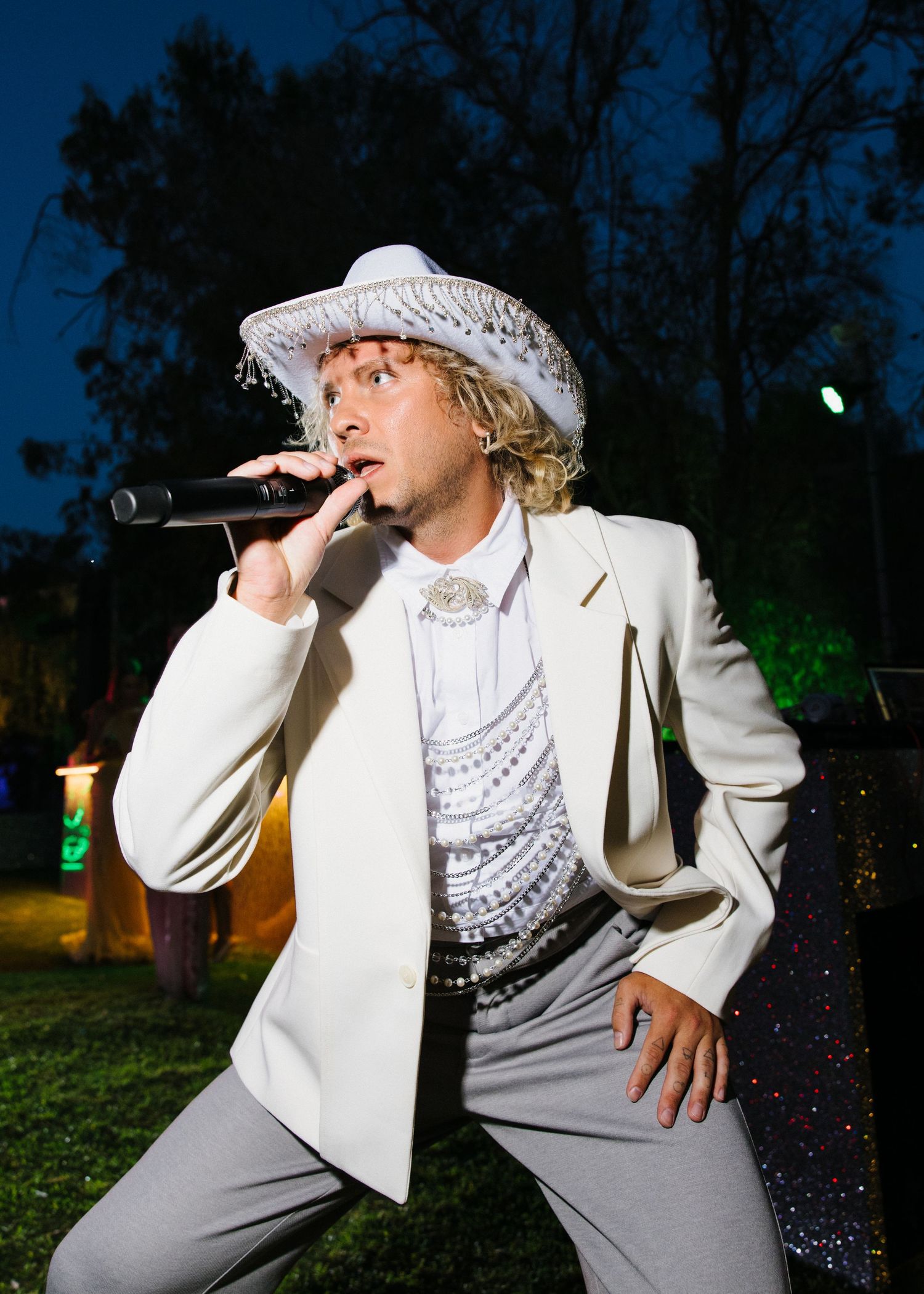 Performer in white suit and lace hat singing into microphone during outdoor event.