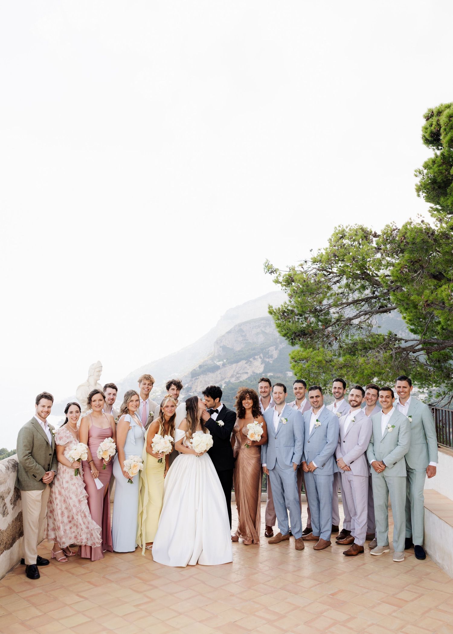 Wedding party poses on a terrace with mountain views, bride in white gown surrounded by bridesmaids and groomsmen.