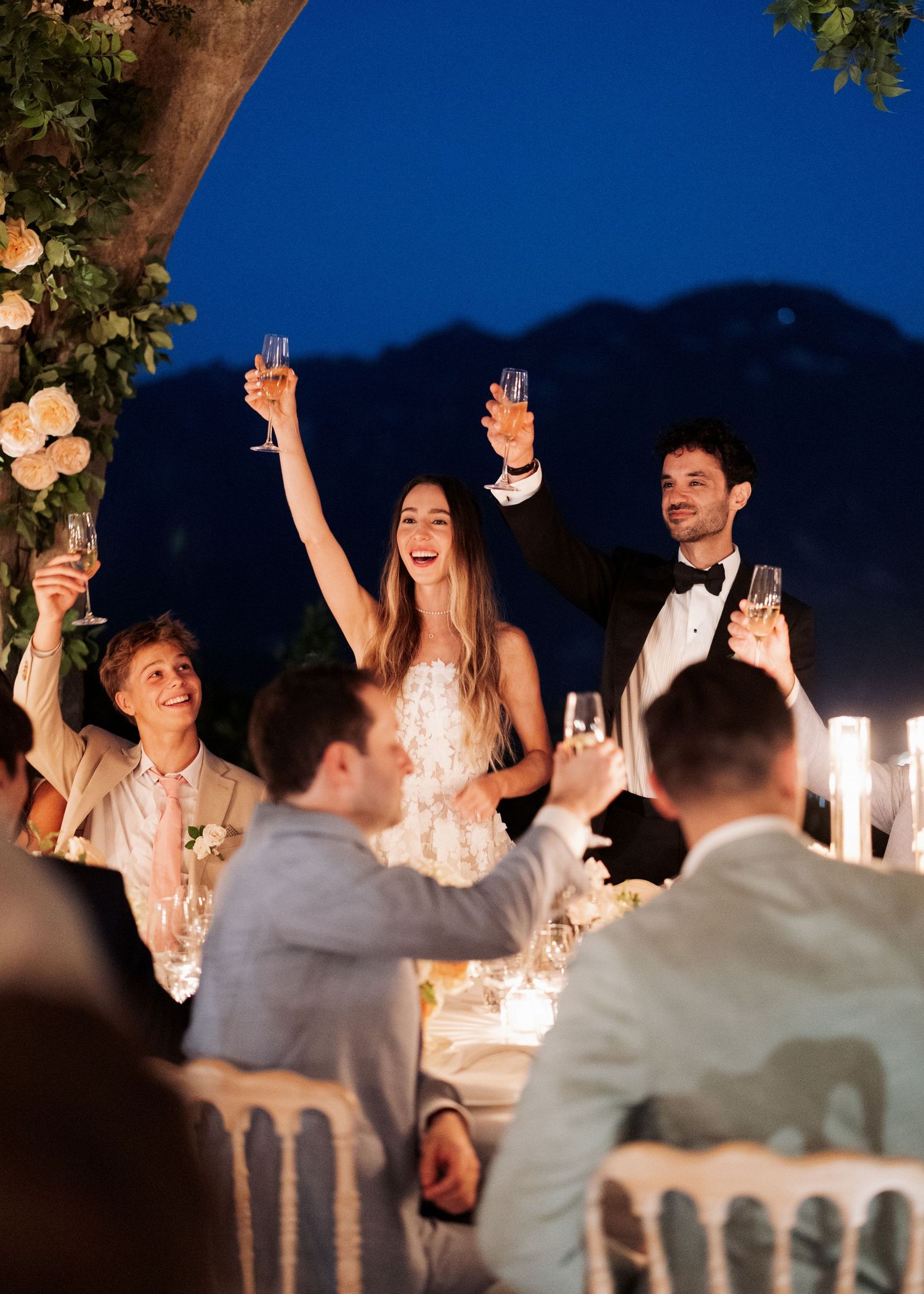 Bride and groom raising champagne glasses during outdoor wedding reception toast at candlelit table under evening sky.