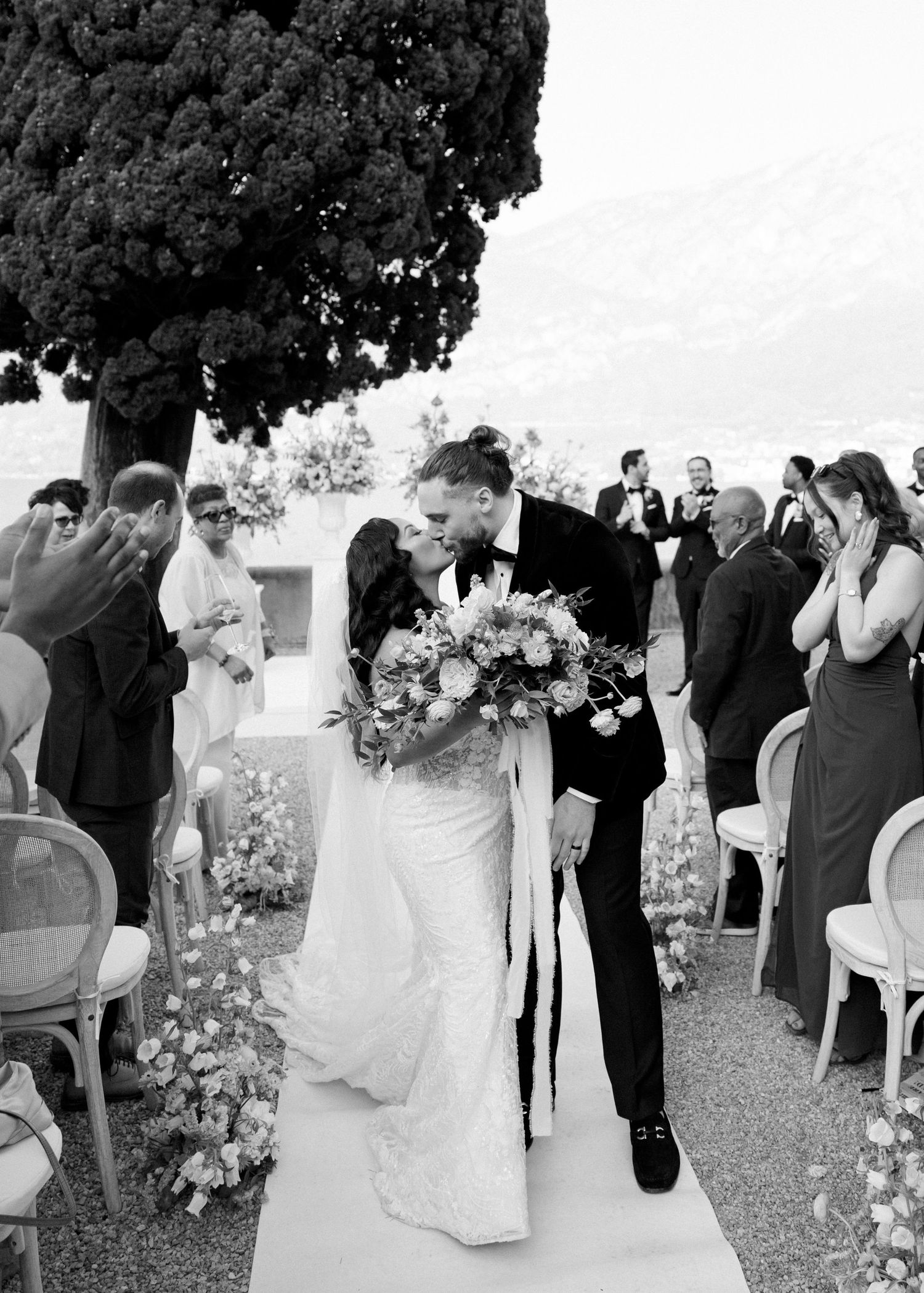 Bride and groom share a kiss at outdoor wedding ceremony aisle, bride holding floral bouquet, guests applauding, black and white.