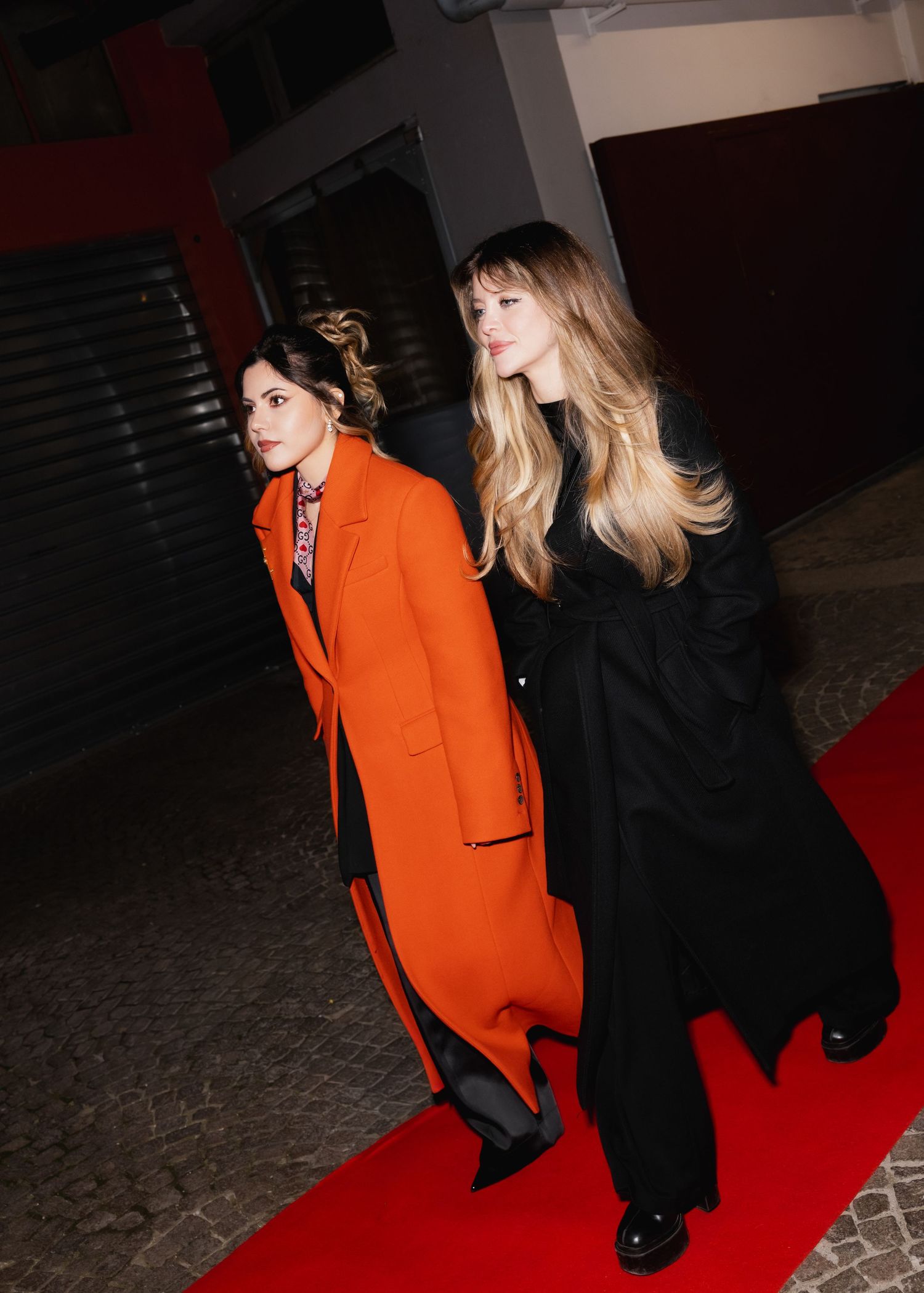 Two models walk the red carpet, one in a striking orange coat and another in black outerwear.