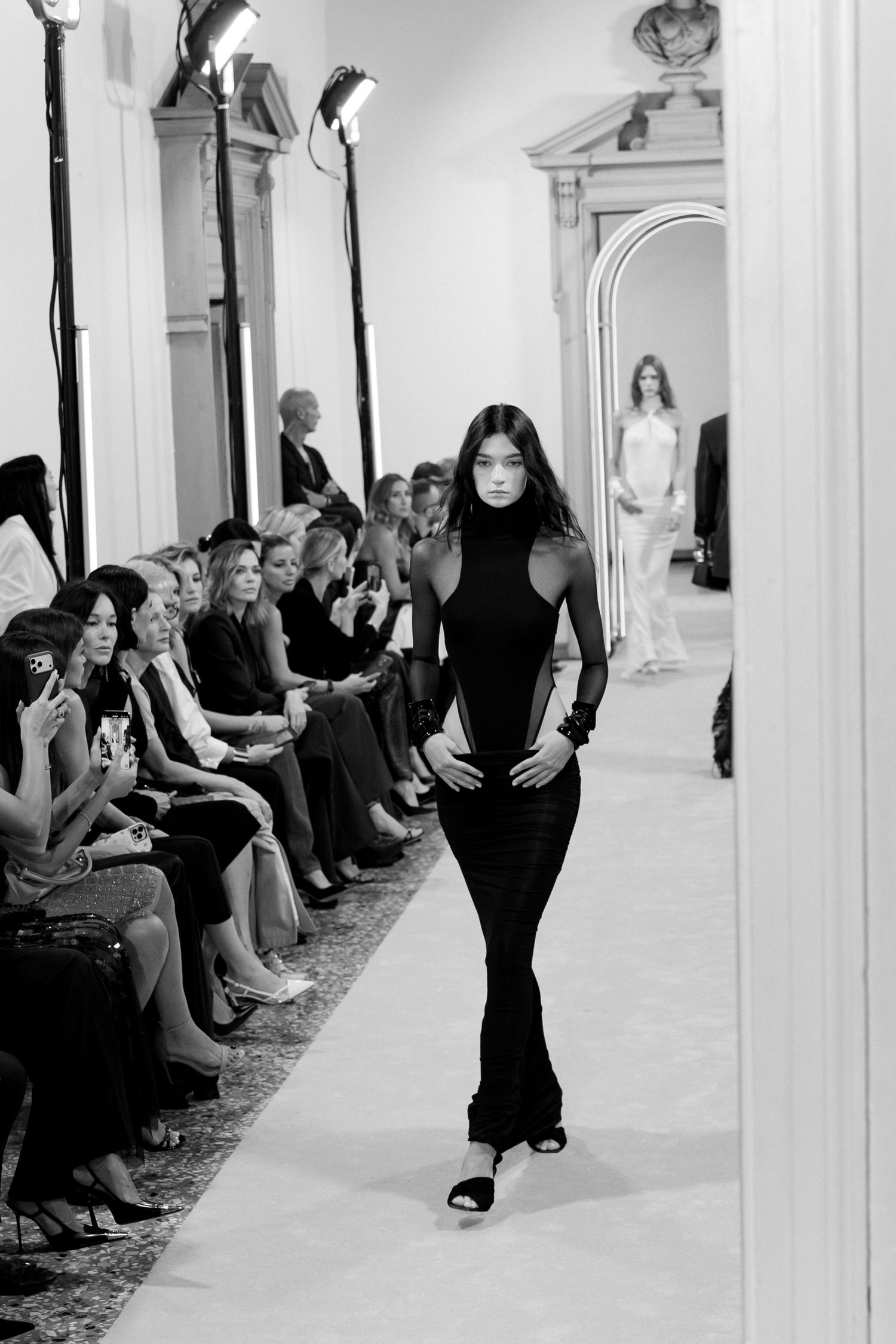 Models walk down a minimalist white runway with audience members seated along the sides during a fashion show.