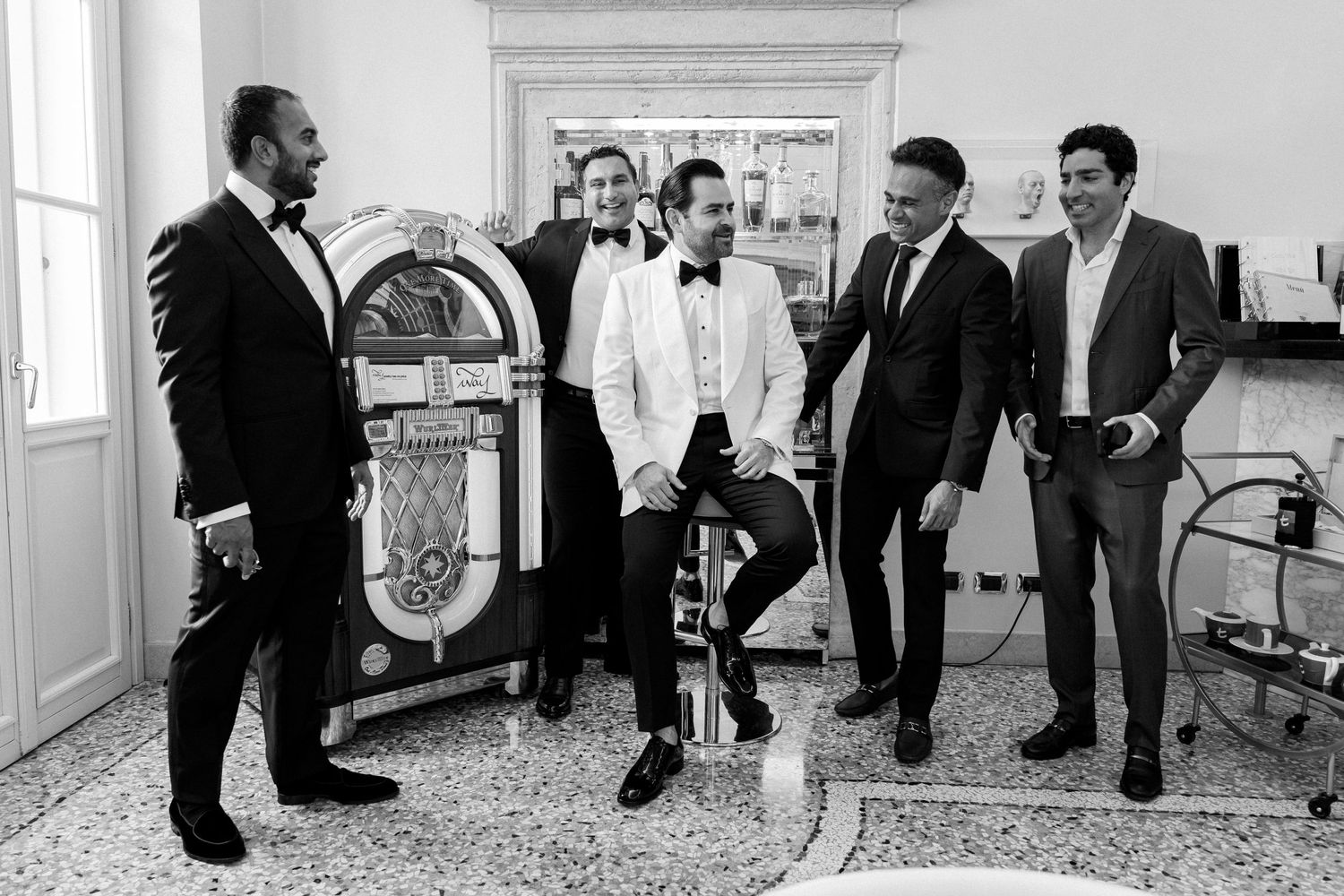 Group of men in formal attire gathered around a vintage jukebox in an elegant room.