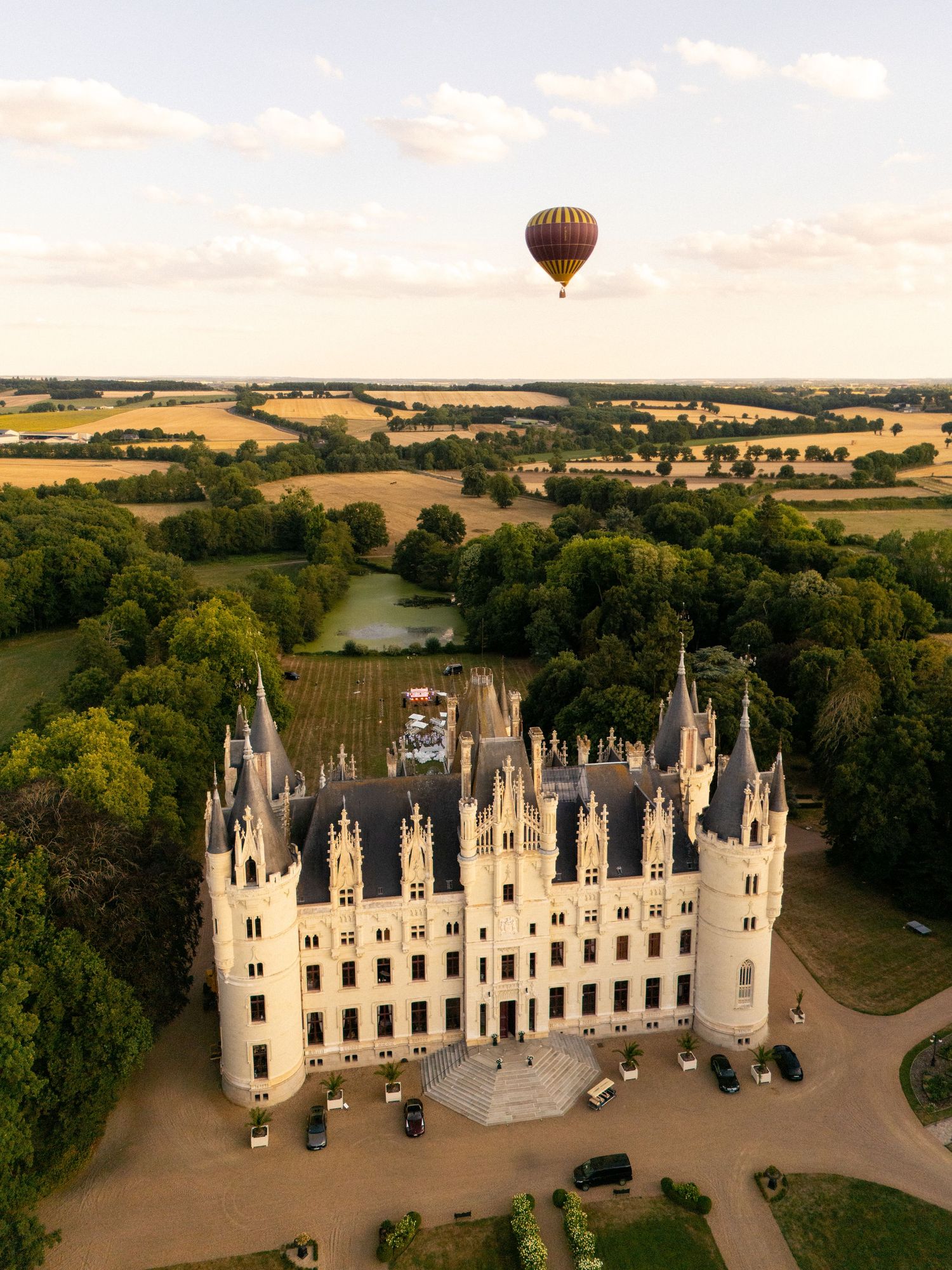 Hot air balloon floats over a majestic French chateau surrounded by lush countryside during golden hour sunset.