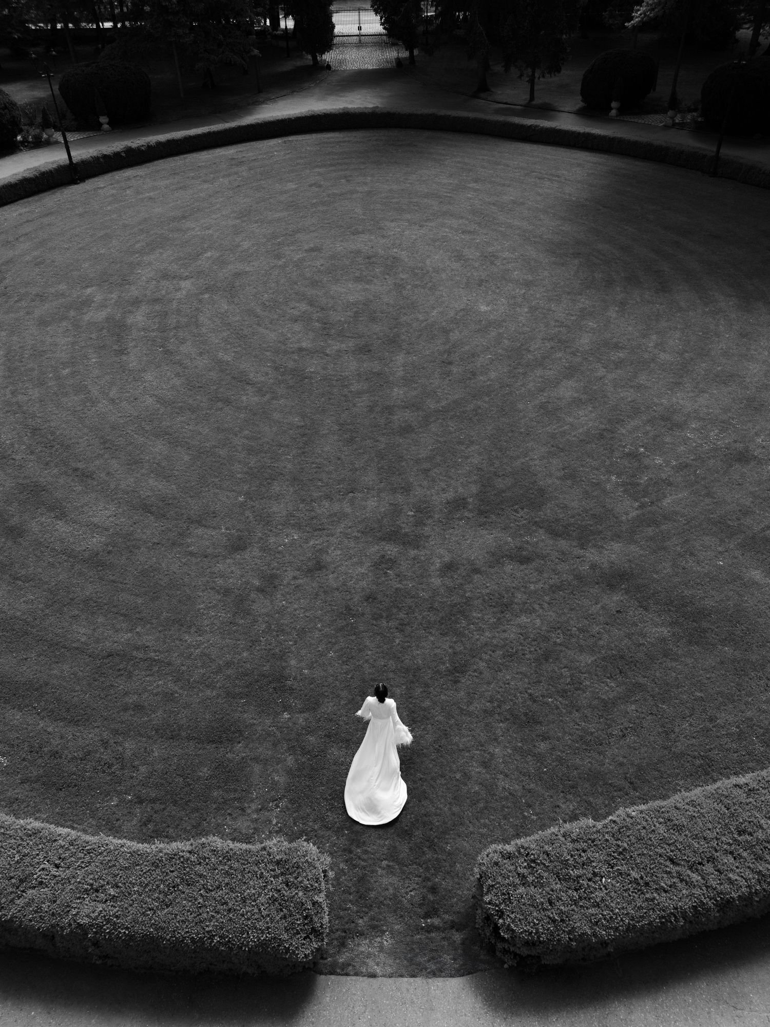 Black and white aerial view of a bride in white dress standing alone on circular manicured lawn garden.