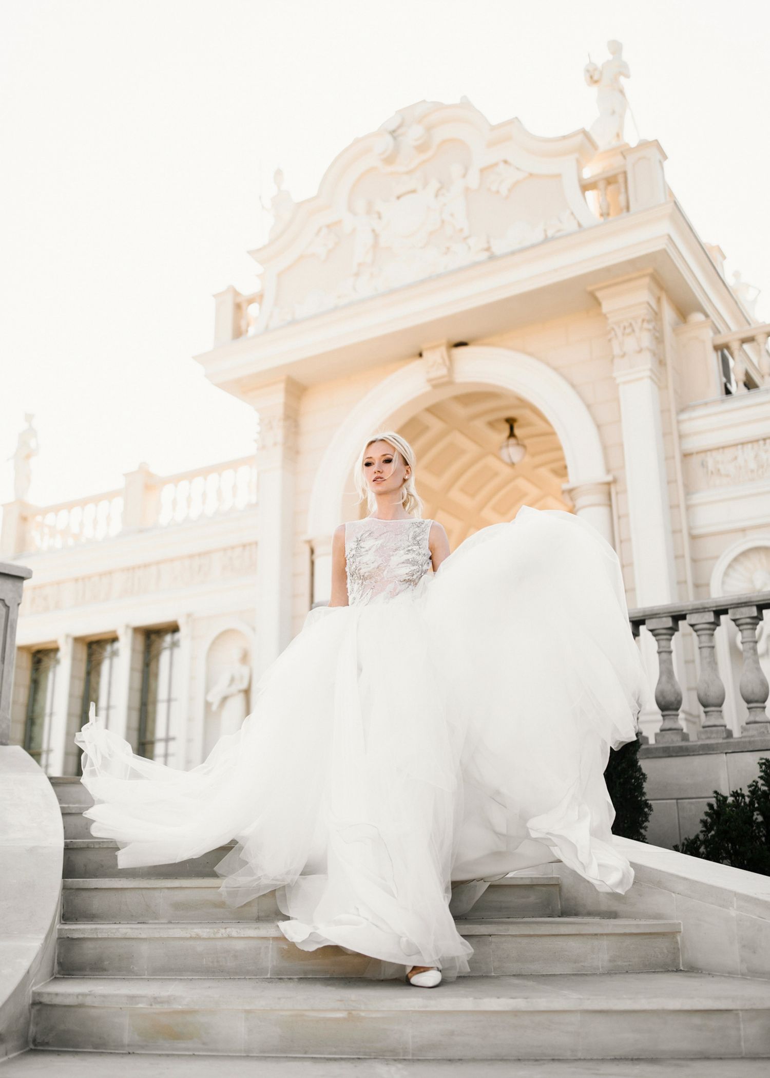 Bride in flowing white gown twirling on marble steps of elegant classical architecture venue.