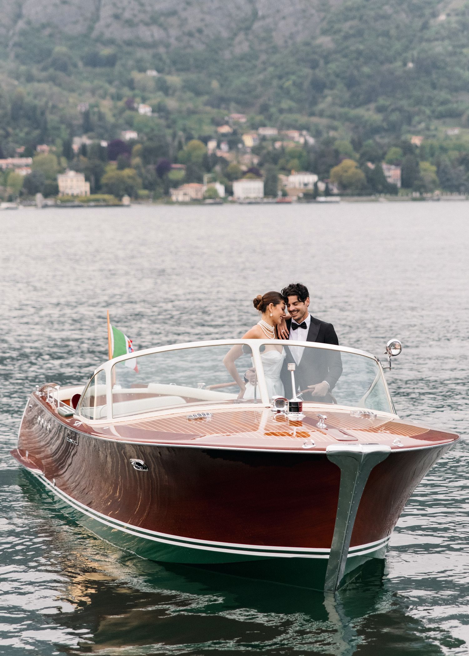 Bride and groom embrace on a classic wooden speedboat on Lake Como, Italy, with lush hillside villas in the background.