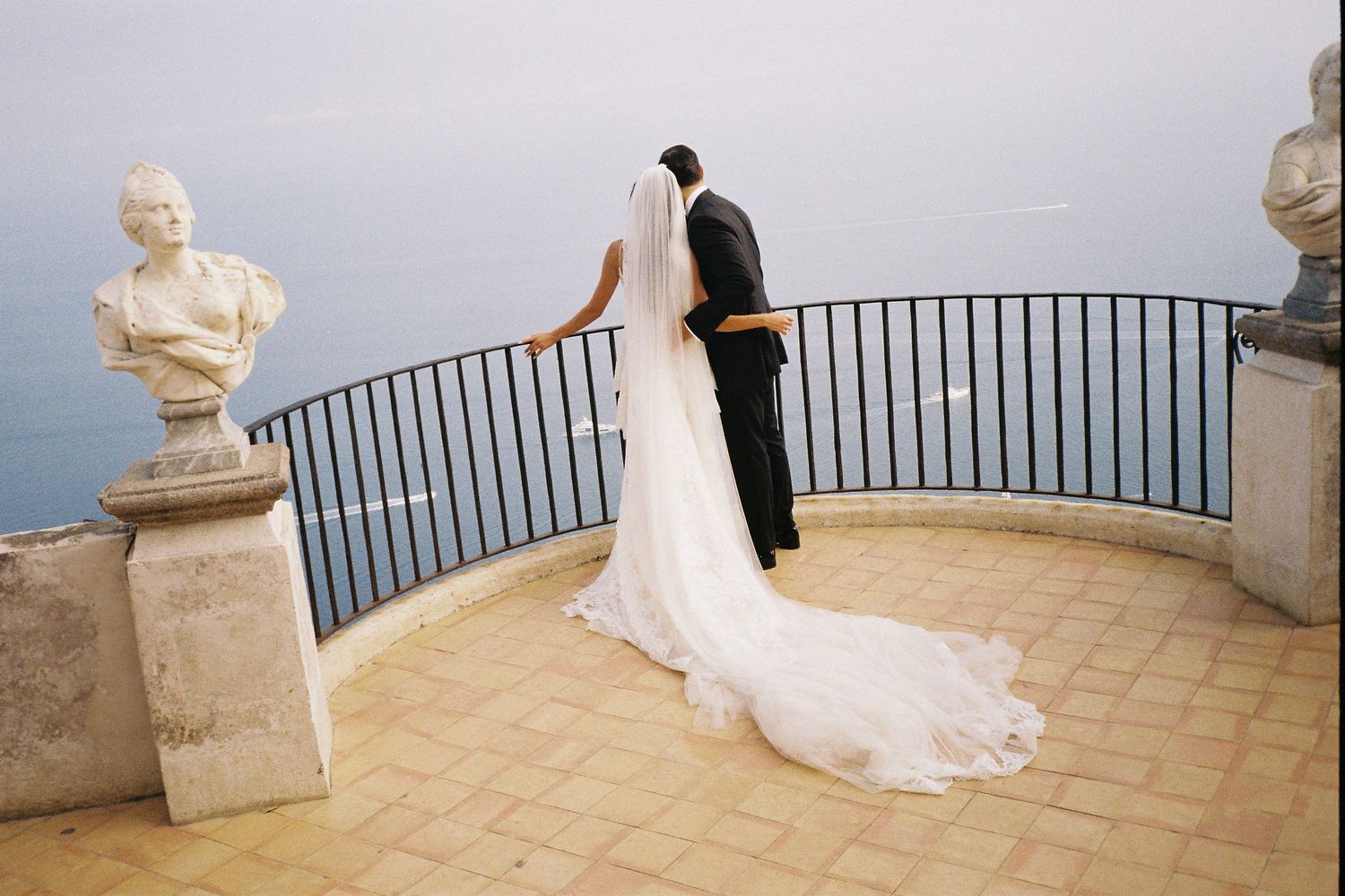 Bride and groom stand at scenic coastal terrace with marble busts, overlooking blue sea at Ravello, Amalfi Coast wedding.
