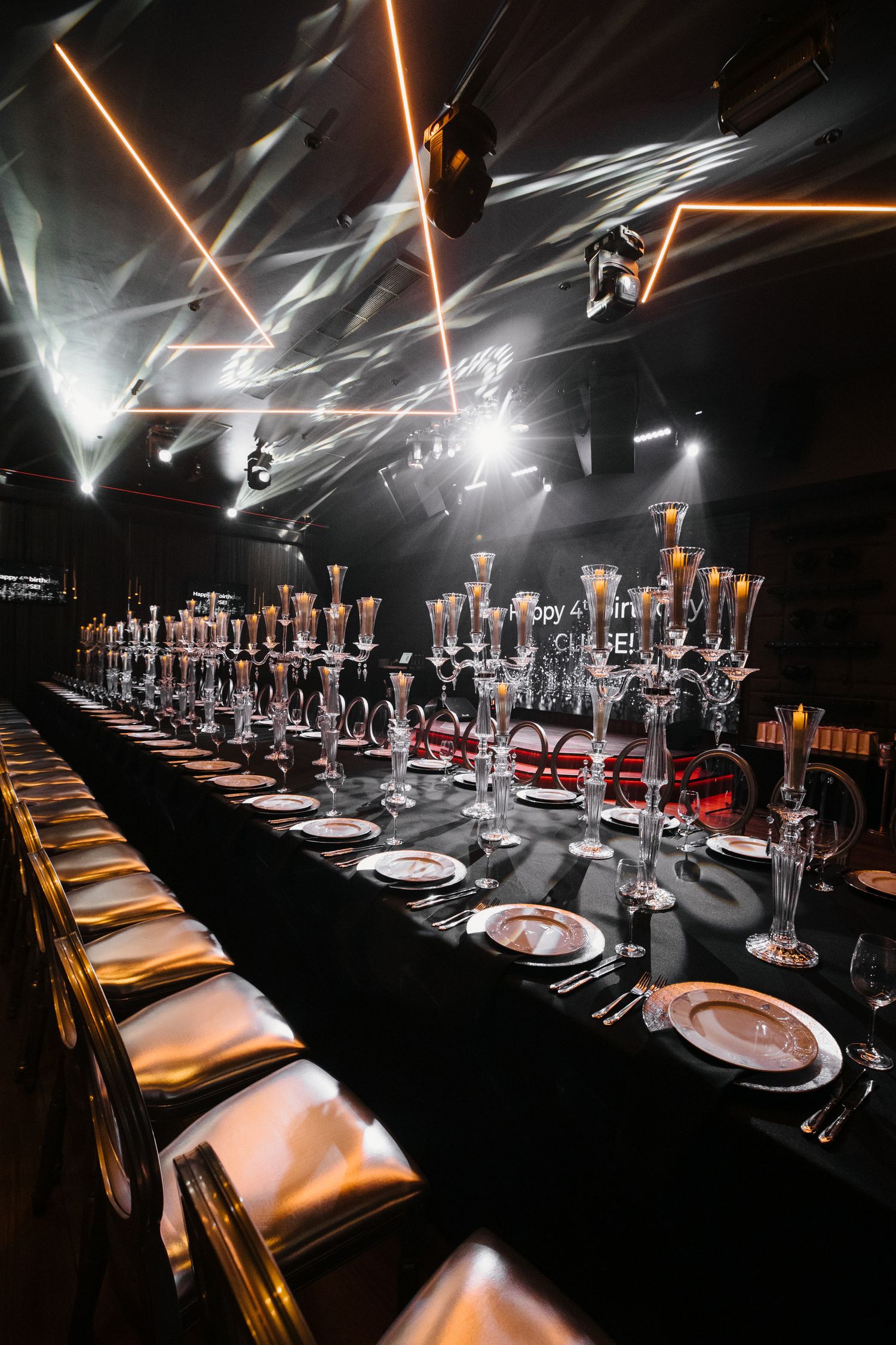 Long formal dining table with dramatic lighting and crystal glassware in dark ambient setting.