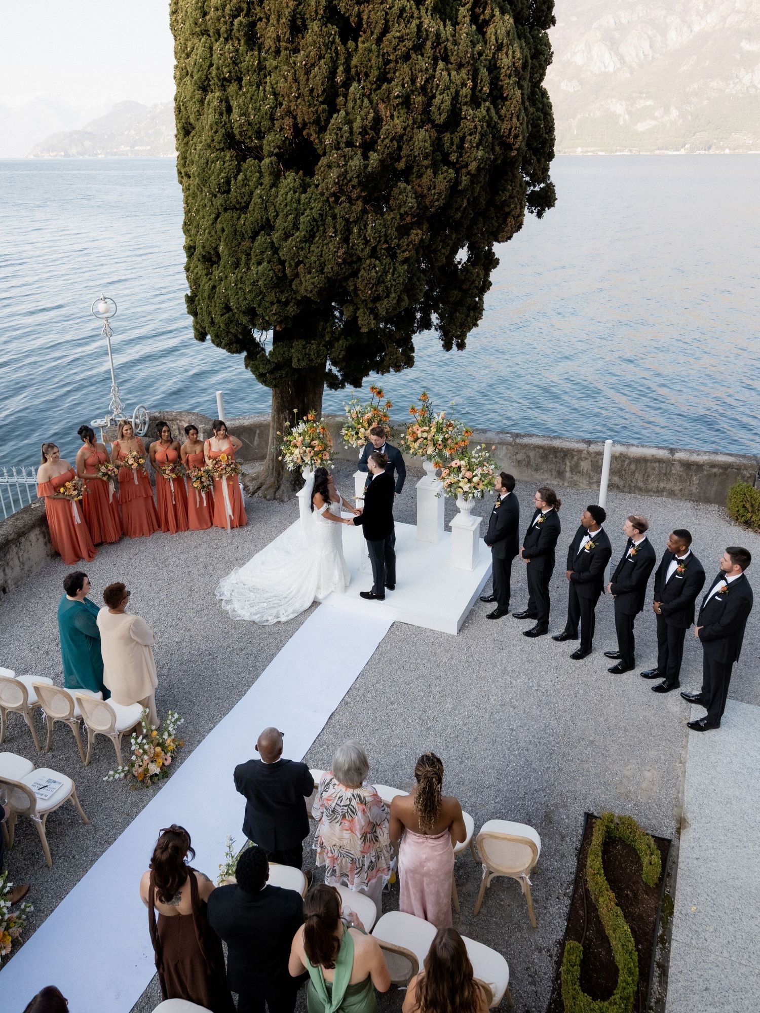 Aerial view of an outdoor wedding ceremony on a lakeside terrace with cypress trees and mountains in the background.