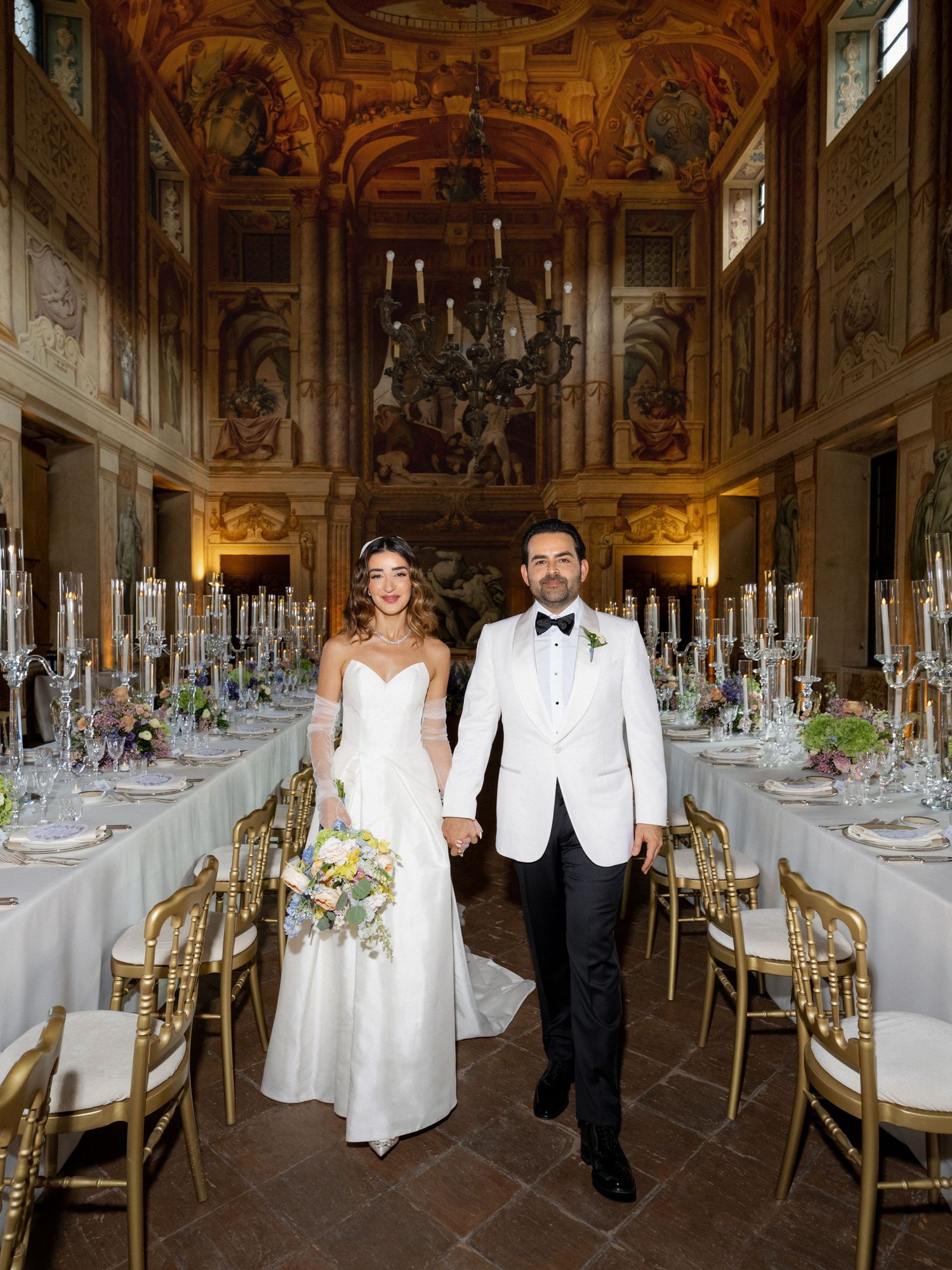 Elegant wedding reception setup in an ornate baroque hall with crystal chandeliers and gold details.