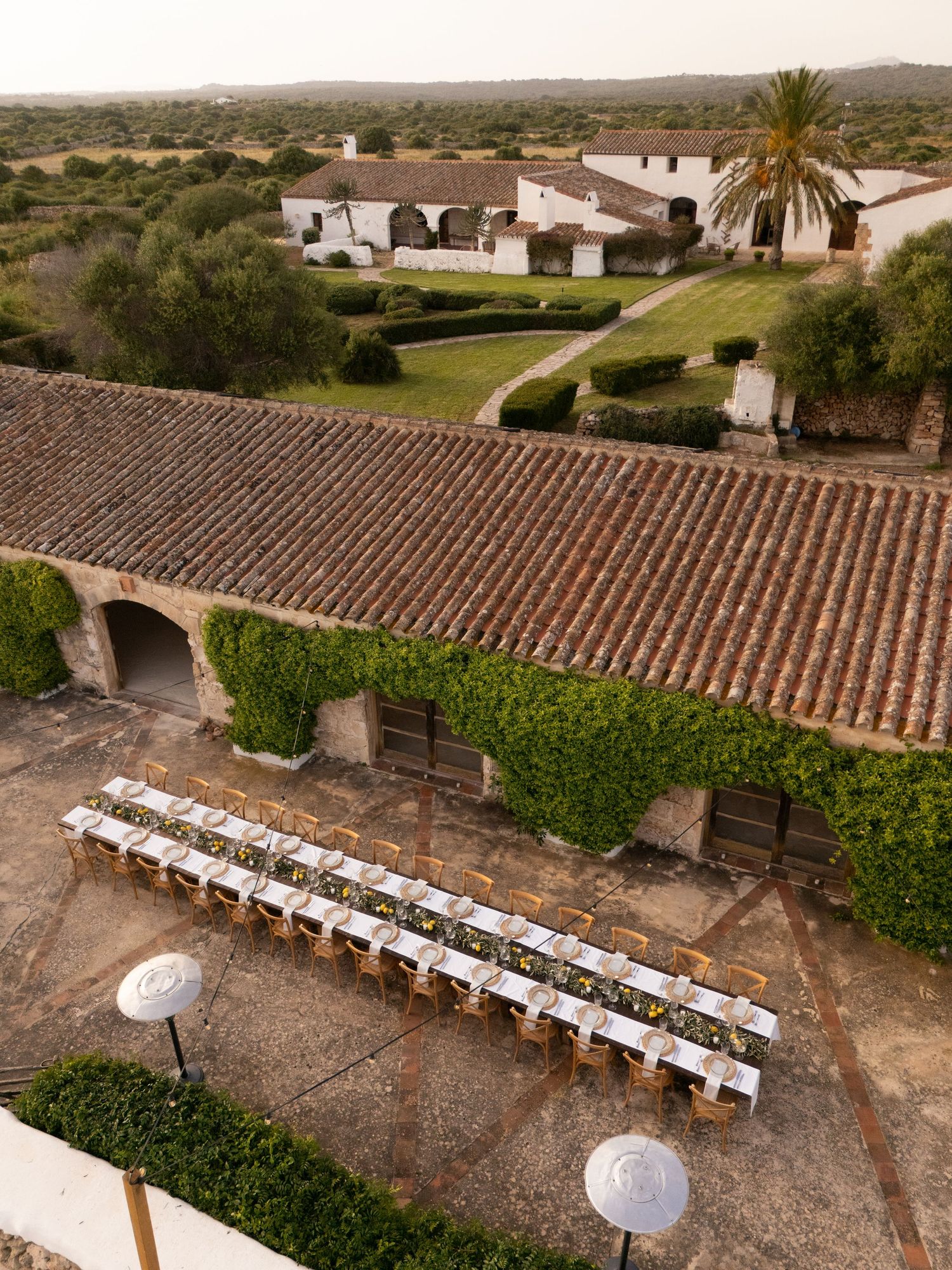 Long banquet tables with white linens are arranged outdoors on a rustic stone terrace overlooking a Mediterranean landscape.