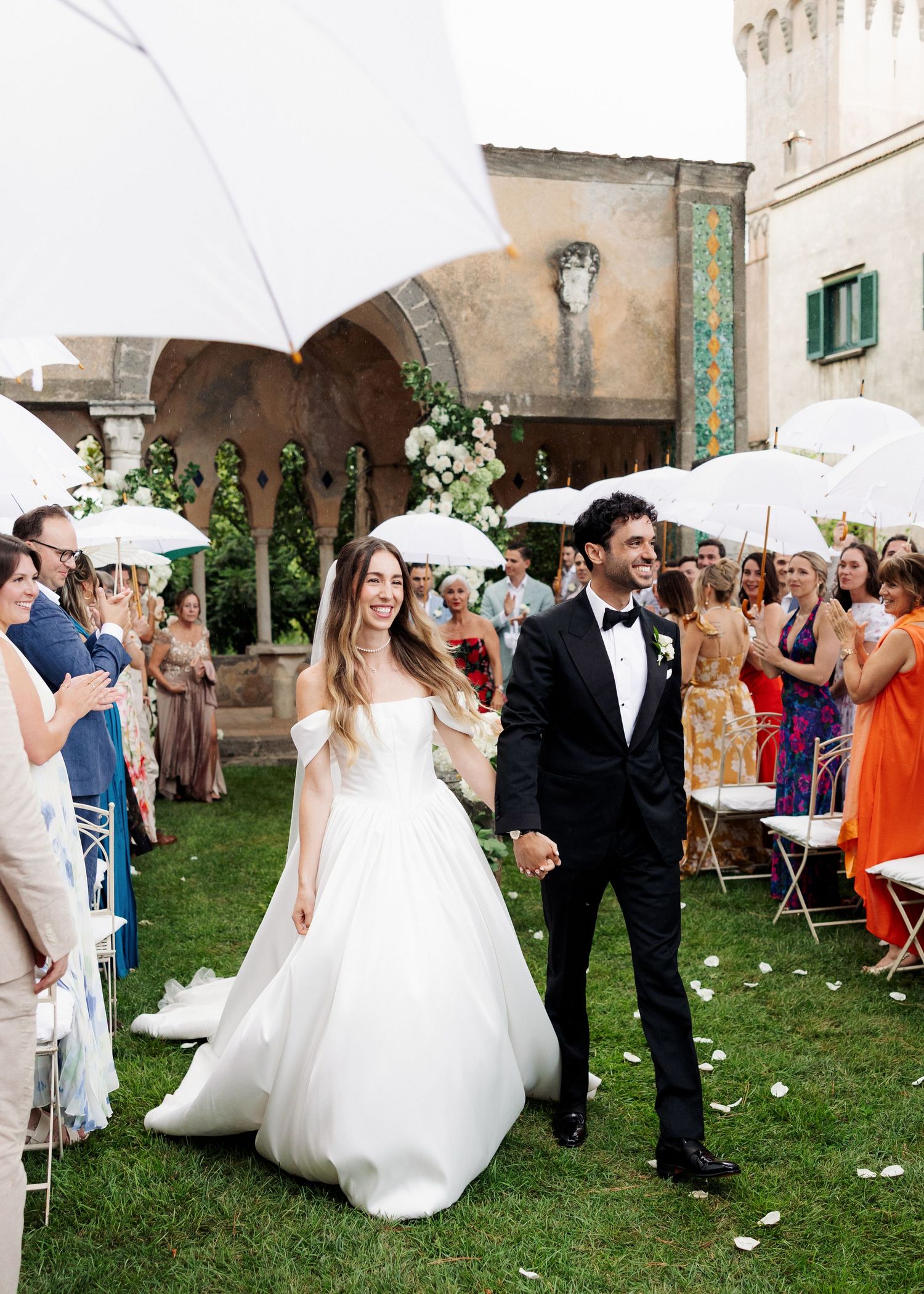Bride and groom walk down outdoor aisle at Italian villa wedding, guests holding white parasols.