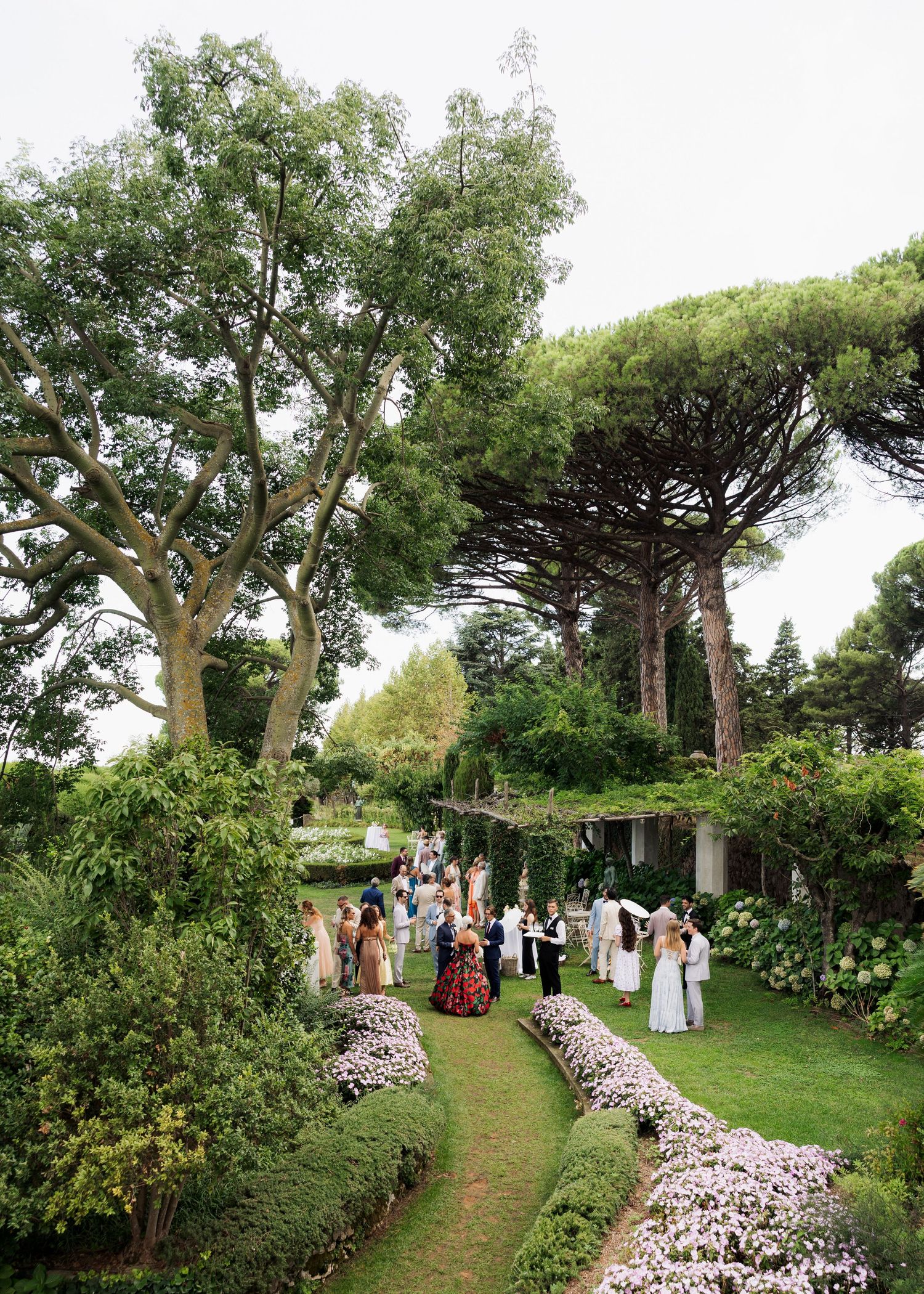 Elegant outdoor garden wedding reception with guests mingling along a lush flower-lined pathway surrounded by tall trees.