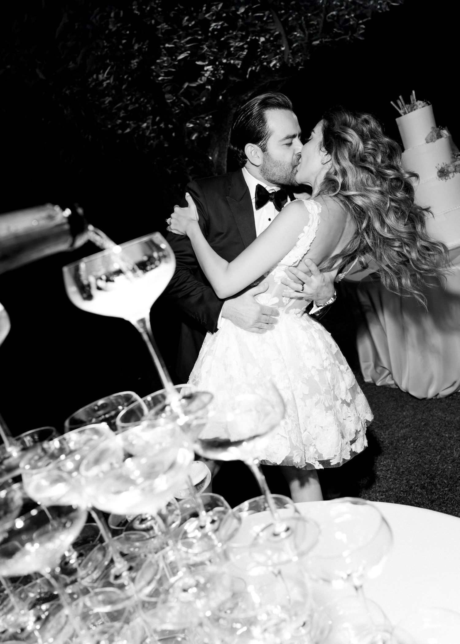 Black and white photo of a bride and groom kissing at their wedding reception with a champagne tower in the foreground.
