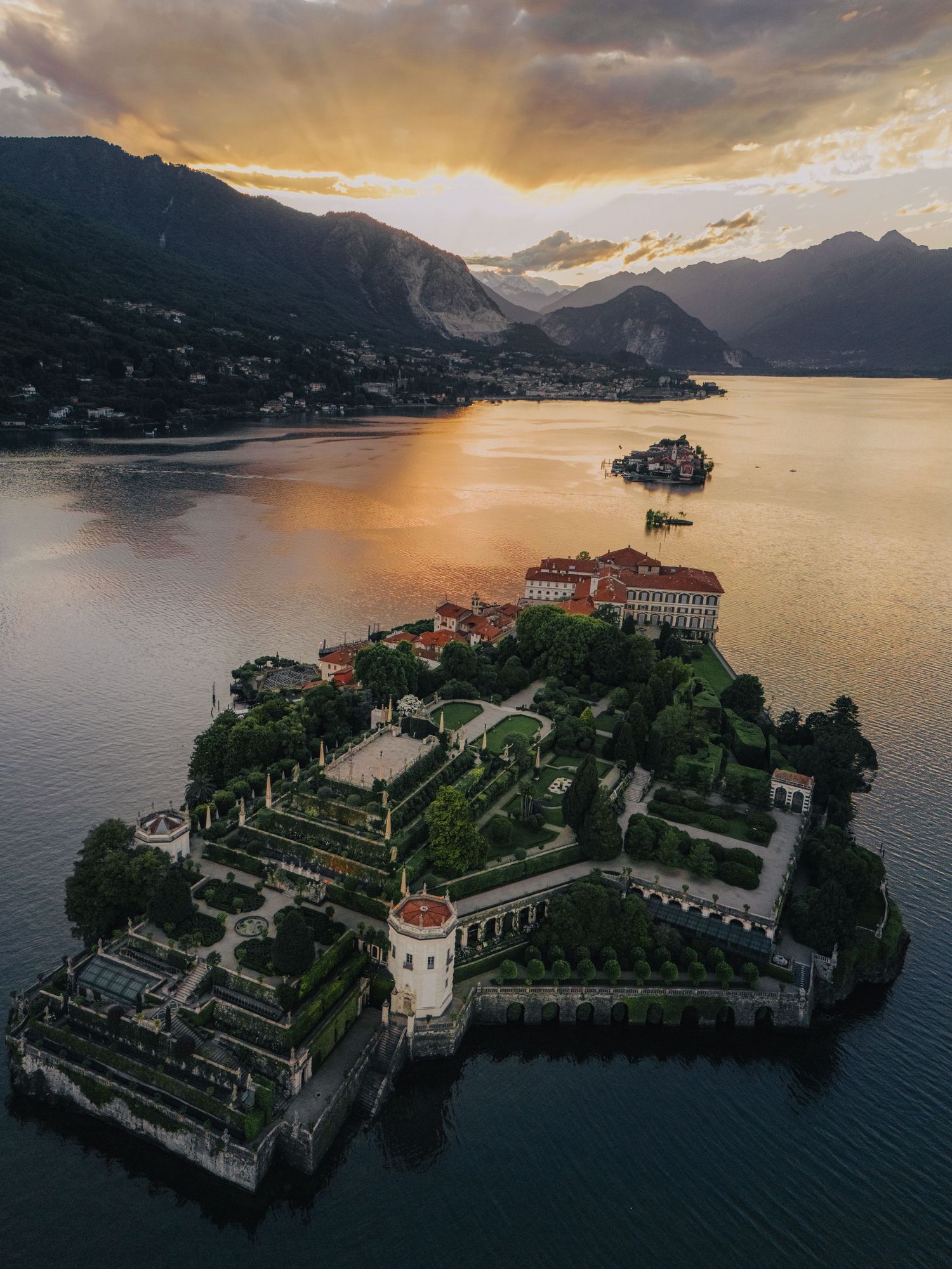 Aerial view of an island palace on Lake Como at sunset with dramatic mountain backdrop.