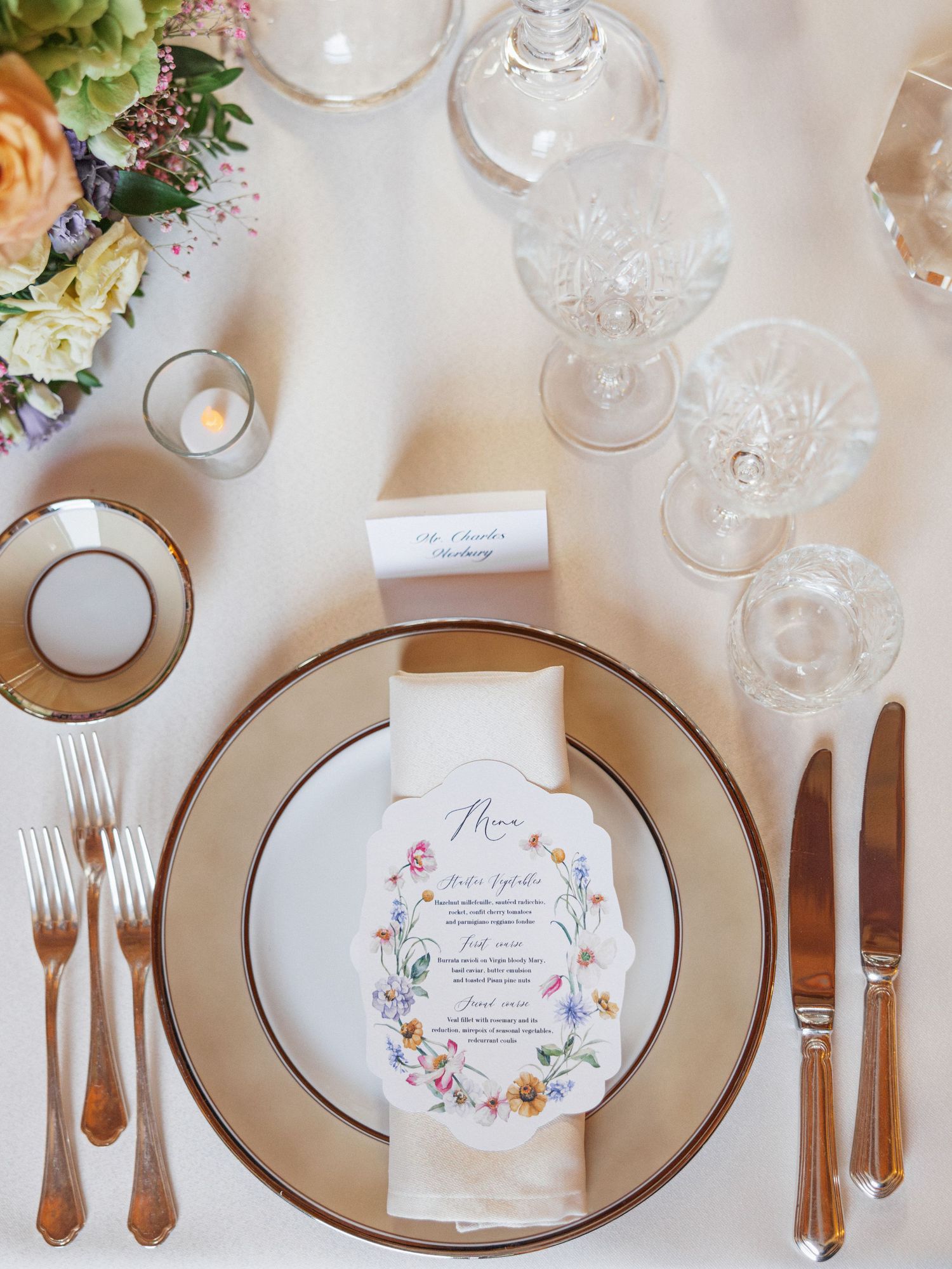 Elegant place setting with floral menu card, crystal glasses and gold-rimmed plates.