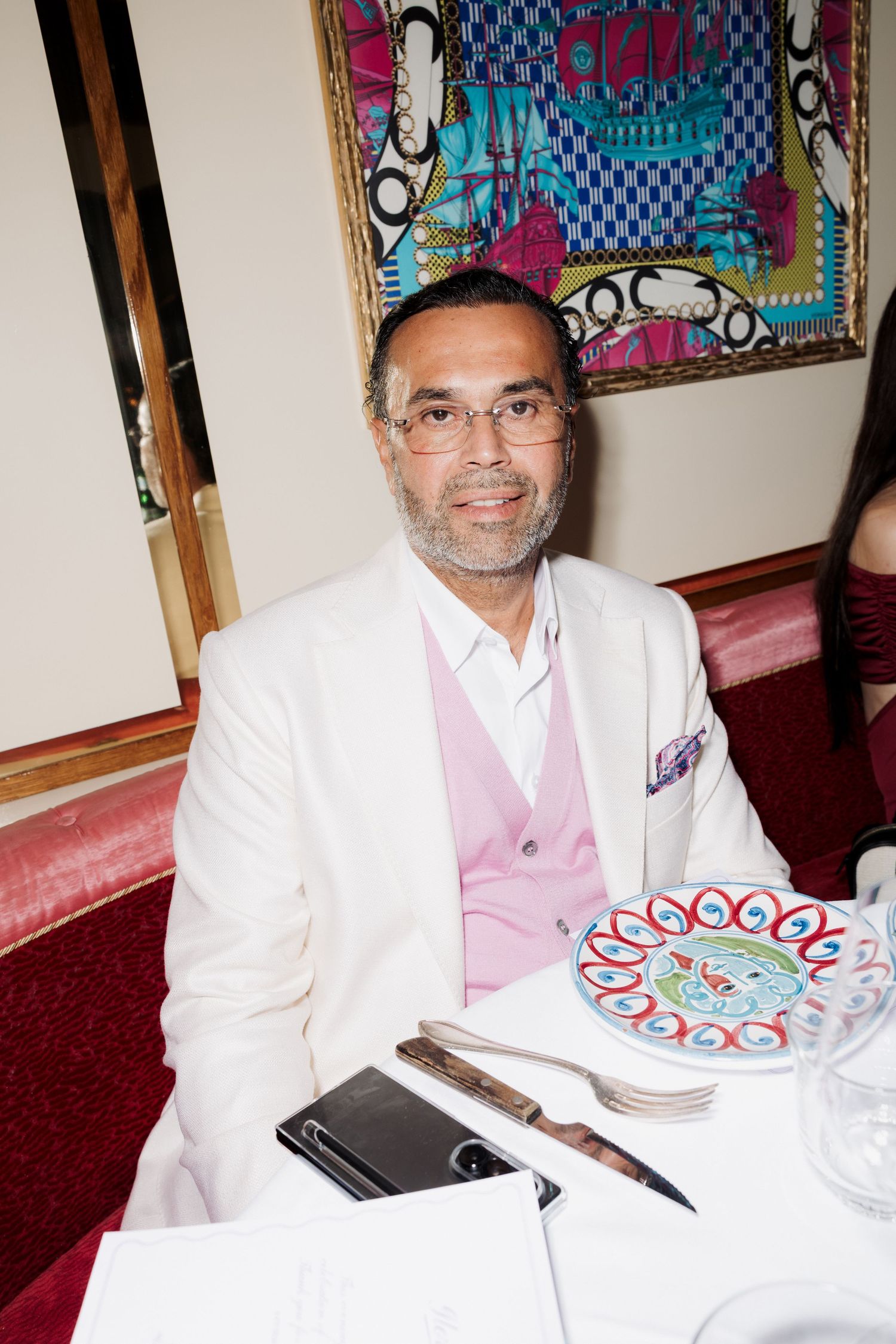 A person in a white suit and pink vest sits at a table with a birthday cake and drinks.